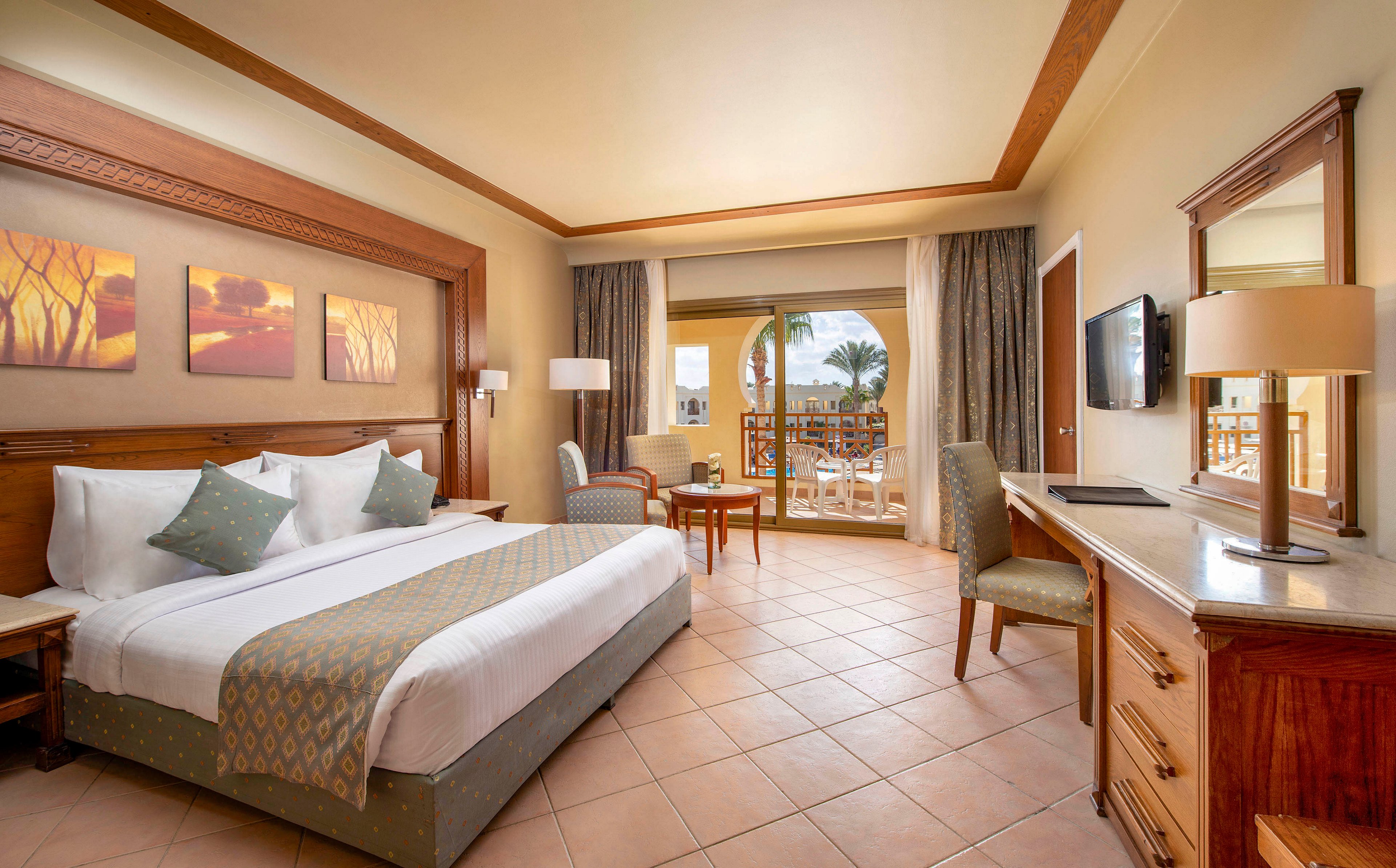 A spacious and well-lit hotel room featuring a large bed with decorative pillows, wooden furniture, and a tiled floor. The room includes a desk, chairs, and a wall-mounted TV, with a balcony offering a scenic outdoor view. Warm tones and natural light create a cozy and inviting atmosphere.