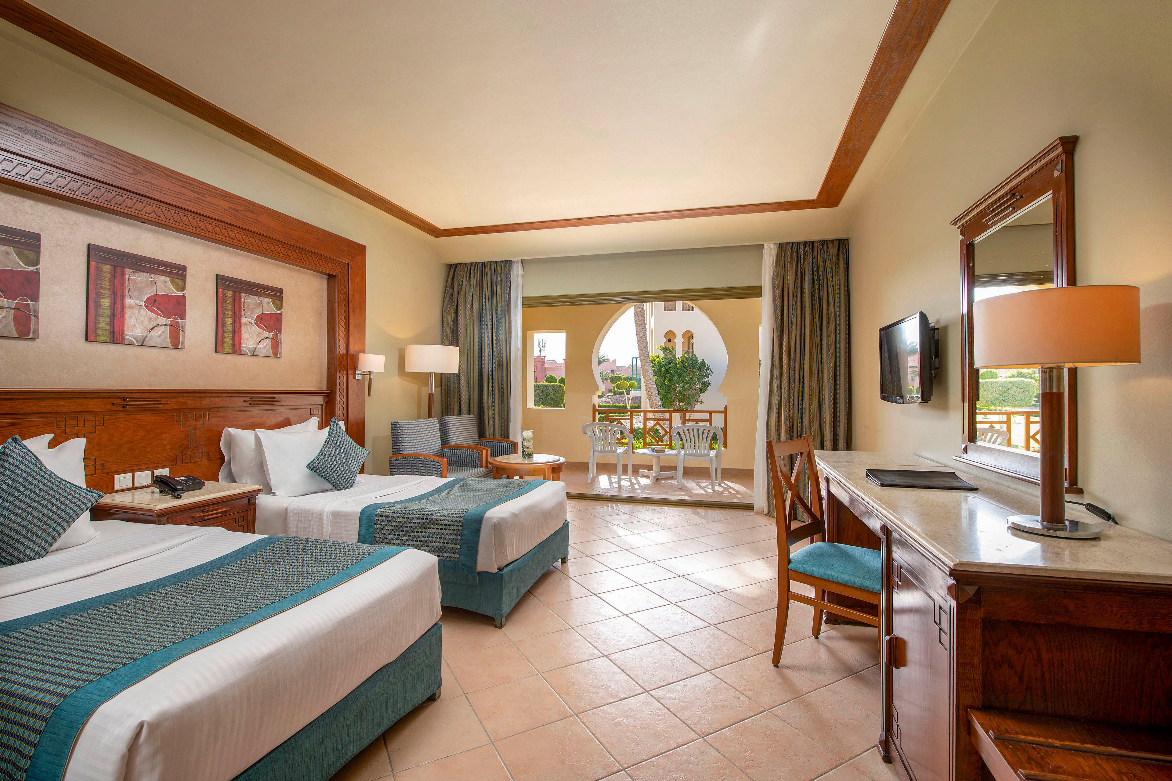 A spacious and well-lit hotel room featuring two neatly made twin beds with teal accents. The room includes a seating area, a desk, and a large mirror, with a balcony overlooking a garden. Warm tones and wooden furniture create a cozy and inviting atmosphere.