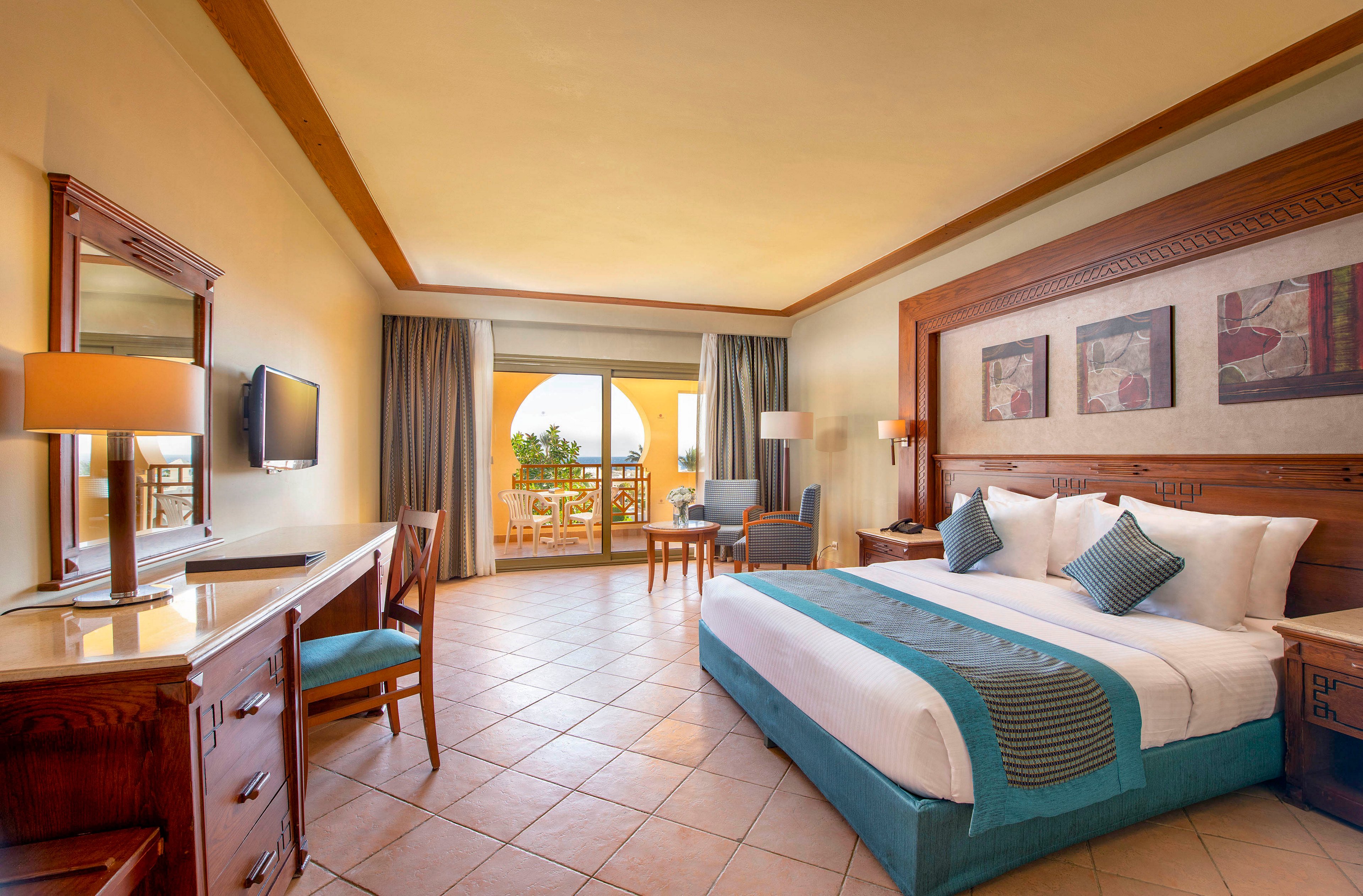A spacious and well-lit hotel room featuring a large bed with teal accents, wooden furniture, and a tiled floor. The room includes a desk, a flat-screen TV, and a seating area near a sliding glass door leading to a balcony with a scenic view. Warm lighting and neutral tones create a cozy and inviting atmosphere.