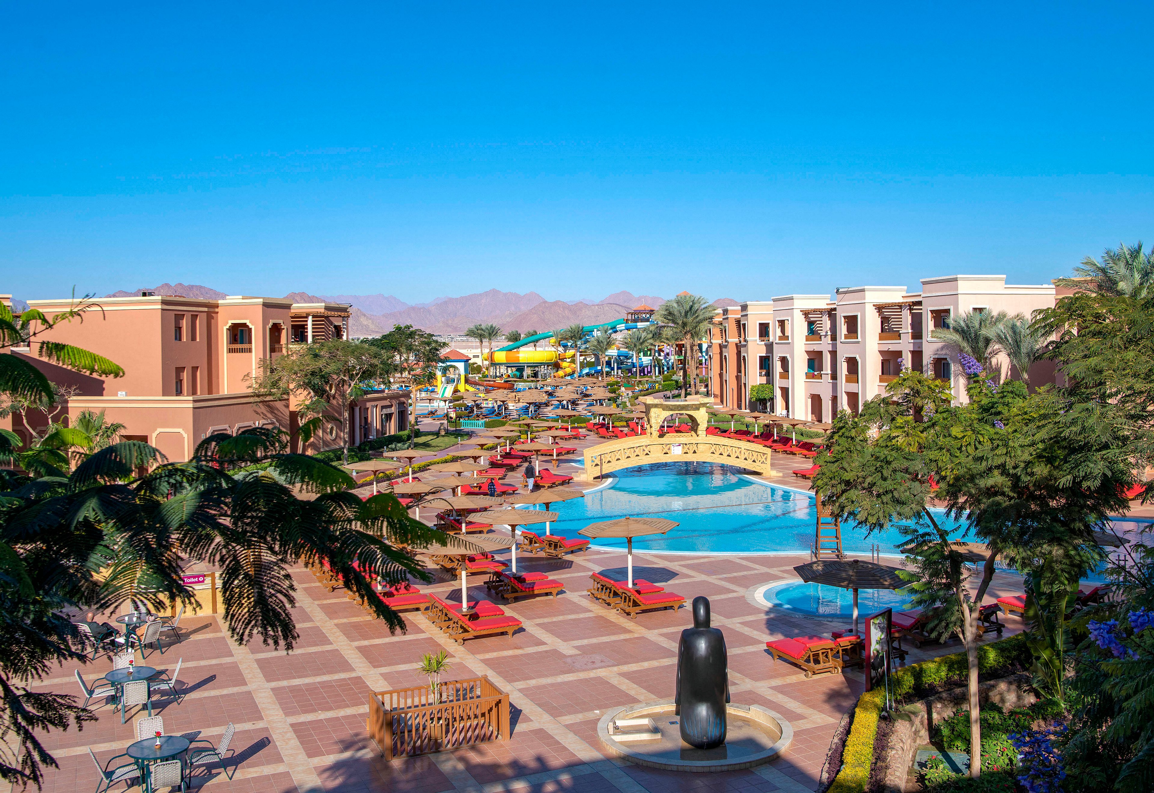 A vibrant resort pool area featuring a large swimming pool surrounded by red lounge chairs and umbrellas. The setting includes palm trees, colorful buildings, and a clear blue sky. A decorative bridge and a statue add to the luxurious ambiance of the space.