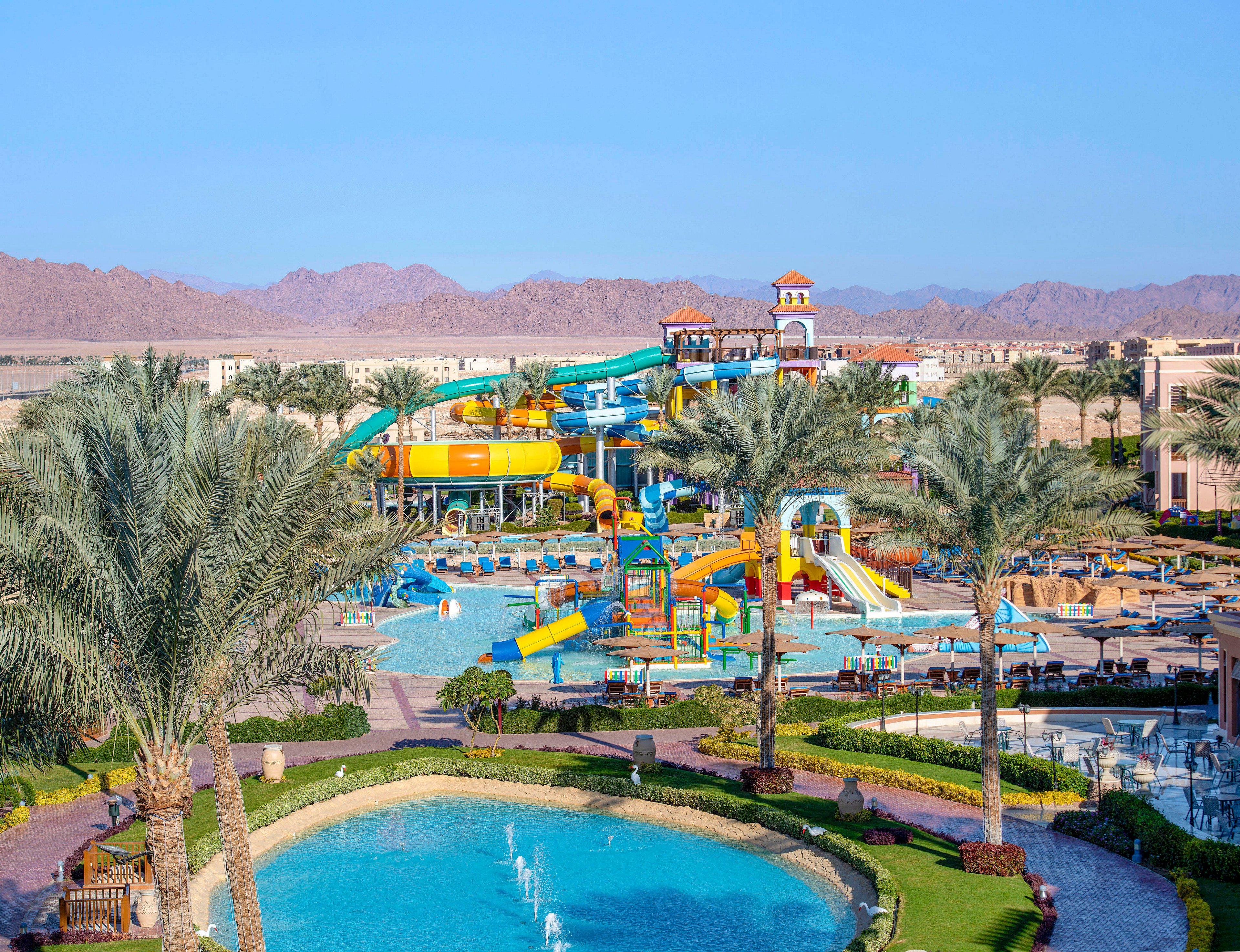 A colorful water park featuring multiple slides, pools, and fountains surrounded by palm trees and landscaped gardens. The setting is sunny with clear blue skies, and desert mountains are visible in the background. The park offers a lively and fun atmosphere for visitors of all ages.
