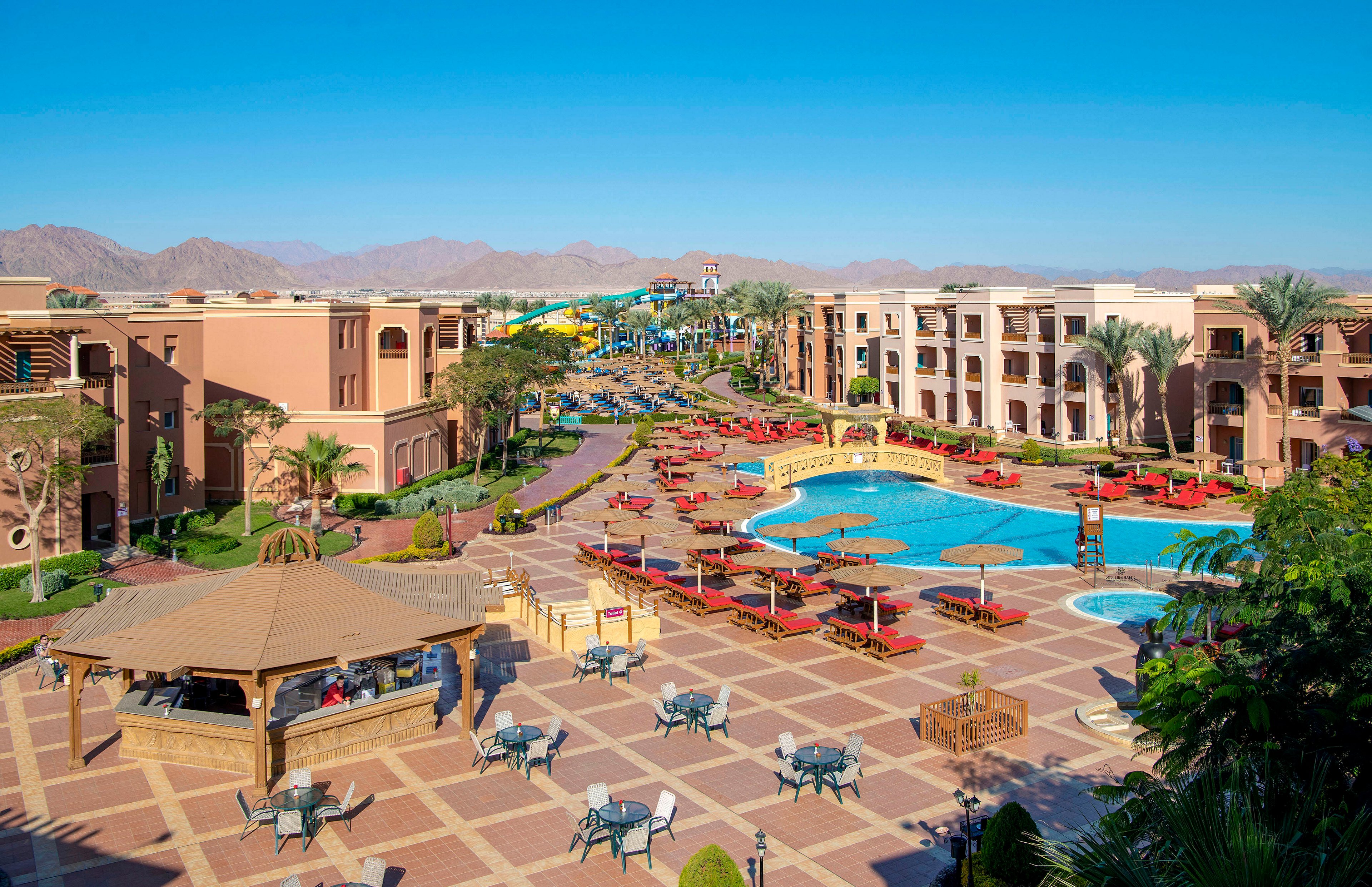 A vibrant luxury resort featuring a large swimming pool surrounded by red sun loungers and umbrellas. The setting includes spacious outdoor seating areas, palm trees, and a clear view of distant mountains under a bright blue sky. The resort's architecture showcases modern design with warm tones, creating a relaxing and inviting atmosphere.