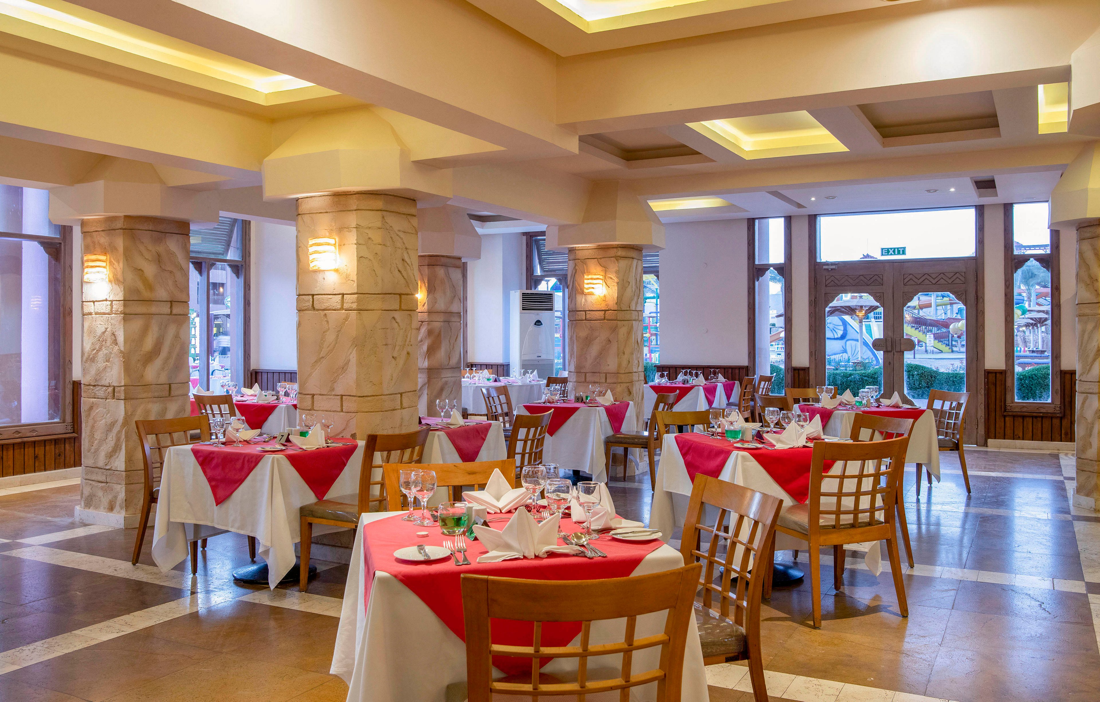 A spacious restaurant dining area featuring neatly arranged tables with red and white tablecloths. The setting includes wooden chairs, stone pillars, and large windows allowing natural light. The warm lighting and organized layout create a welcoming and refined atmosphere.