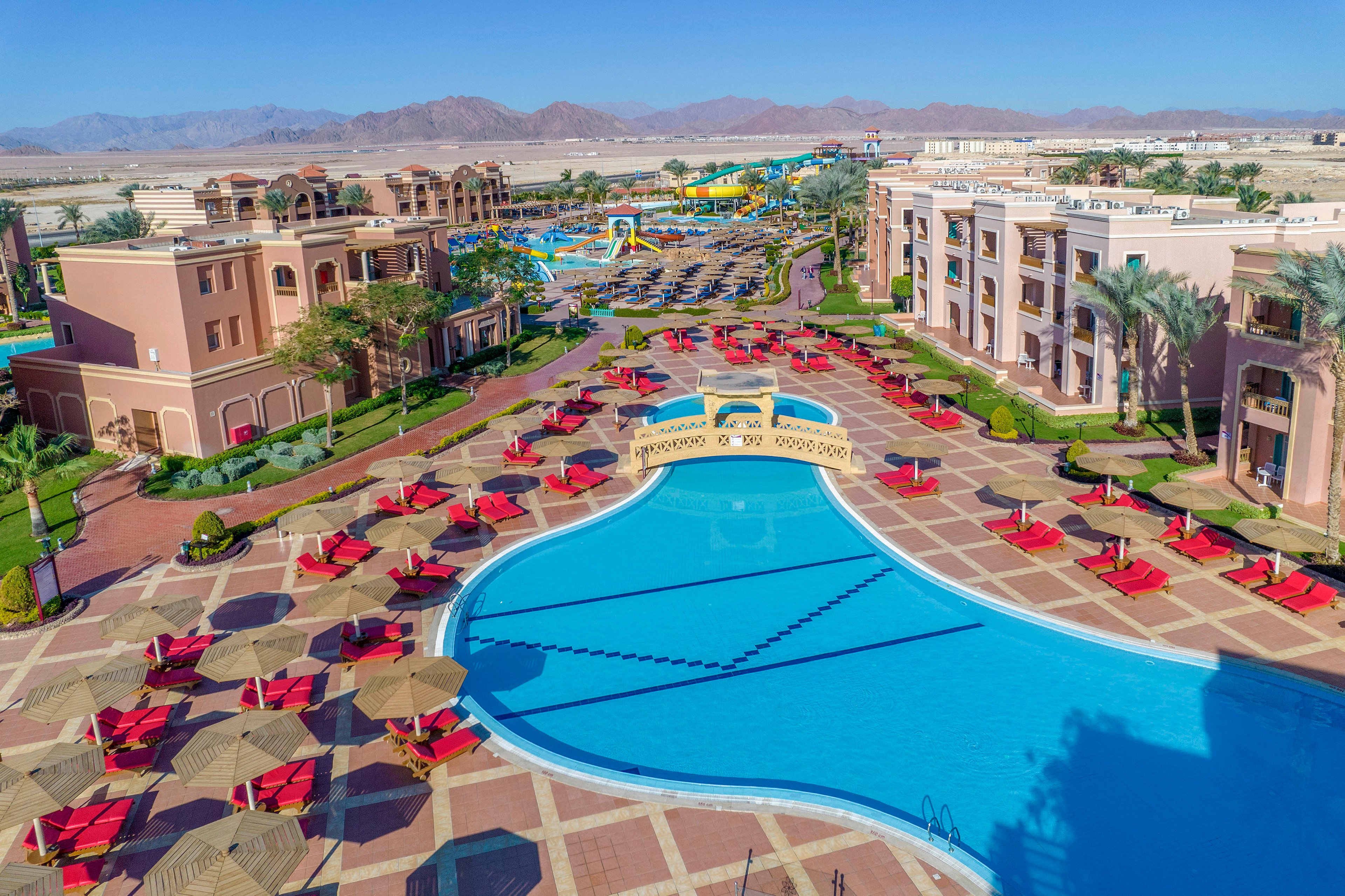 A large resort pool surrounded by red loungers and straw umbrellas in a sunny outdoor setting. The area features tiled walkways and landscaped greenery, with additional water slides and buildings visible in the background. The scene is framed by distant mountains under a clear blue sky.