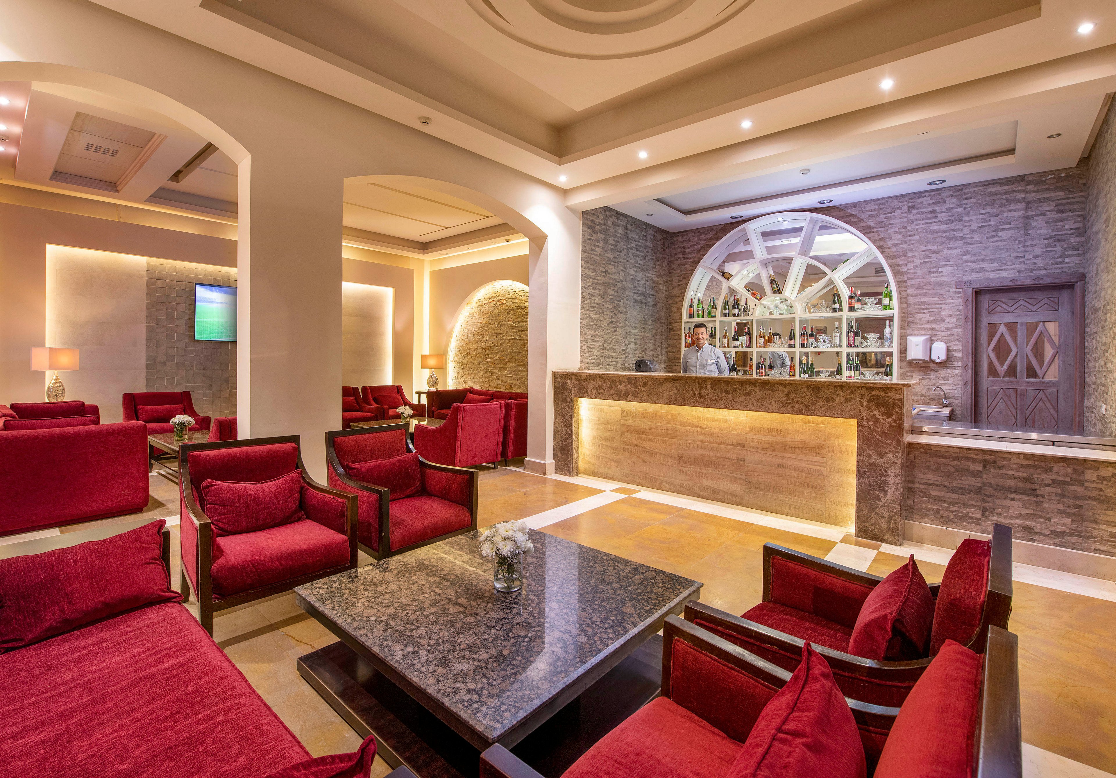 A stylish hotel lounge featuring a modern bar counter with a well-lit display of bottles. The seating area includes plush red armchairs and a central granite coffee table. Warm lighting and arched architectural details create a cozy and inviting atmosphere.