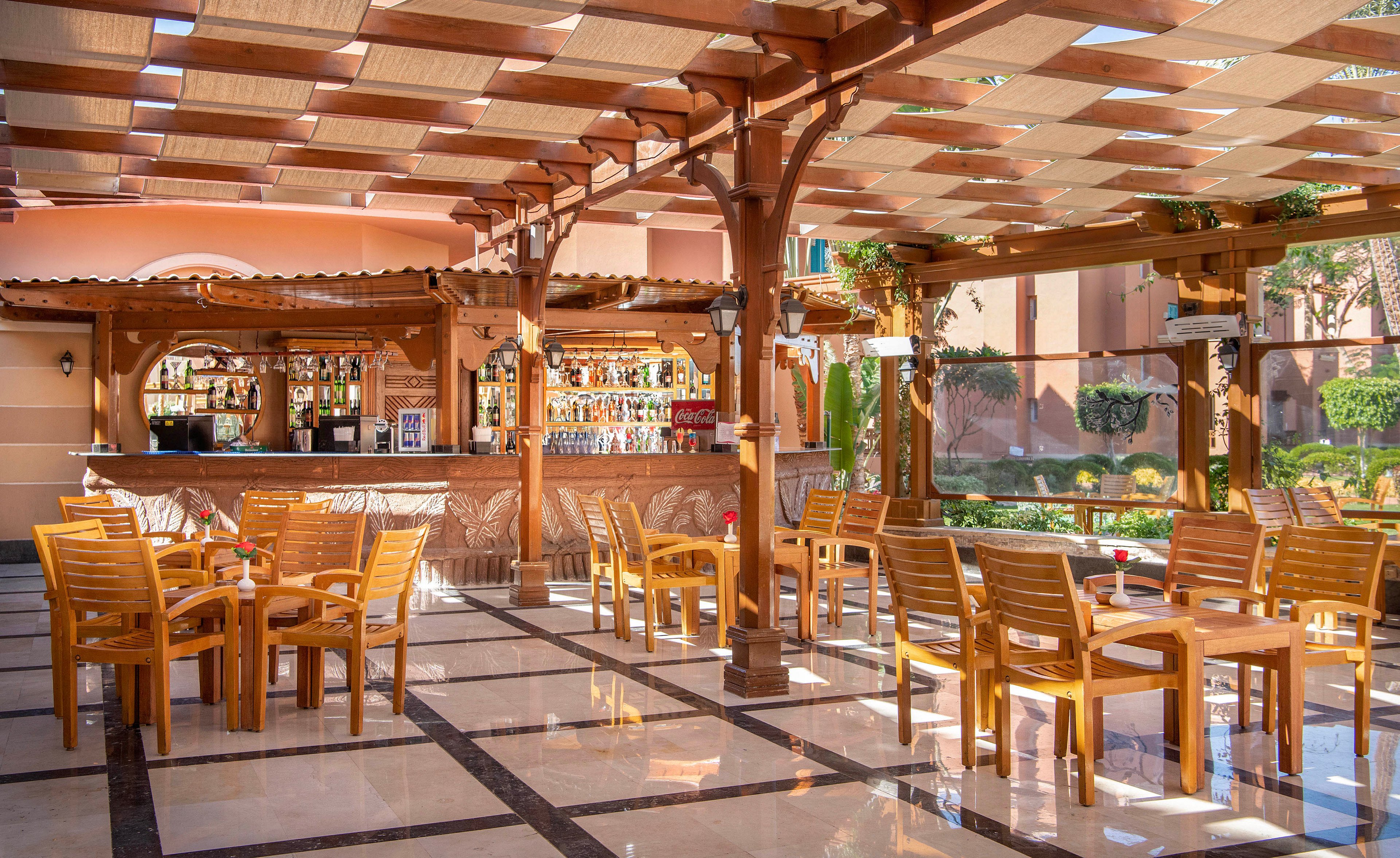 An inviting outdoor bar with wooden chairs and tables under a pergola-style roof. The bar is stocked with various bottles and features a polished countertop. The setting is bright and sunny, with greenery visible in the background, creating a relaxed and welcoming atmosphere.
