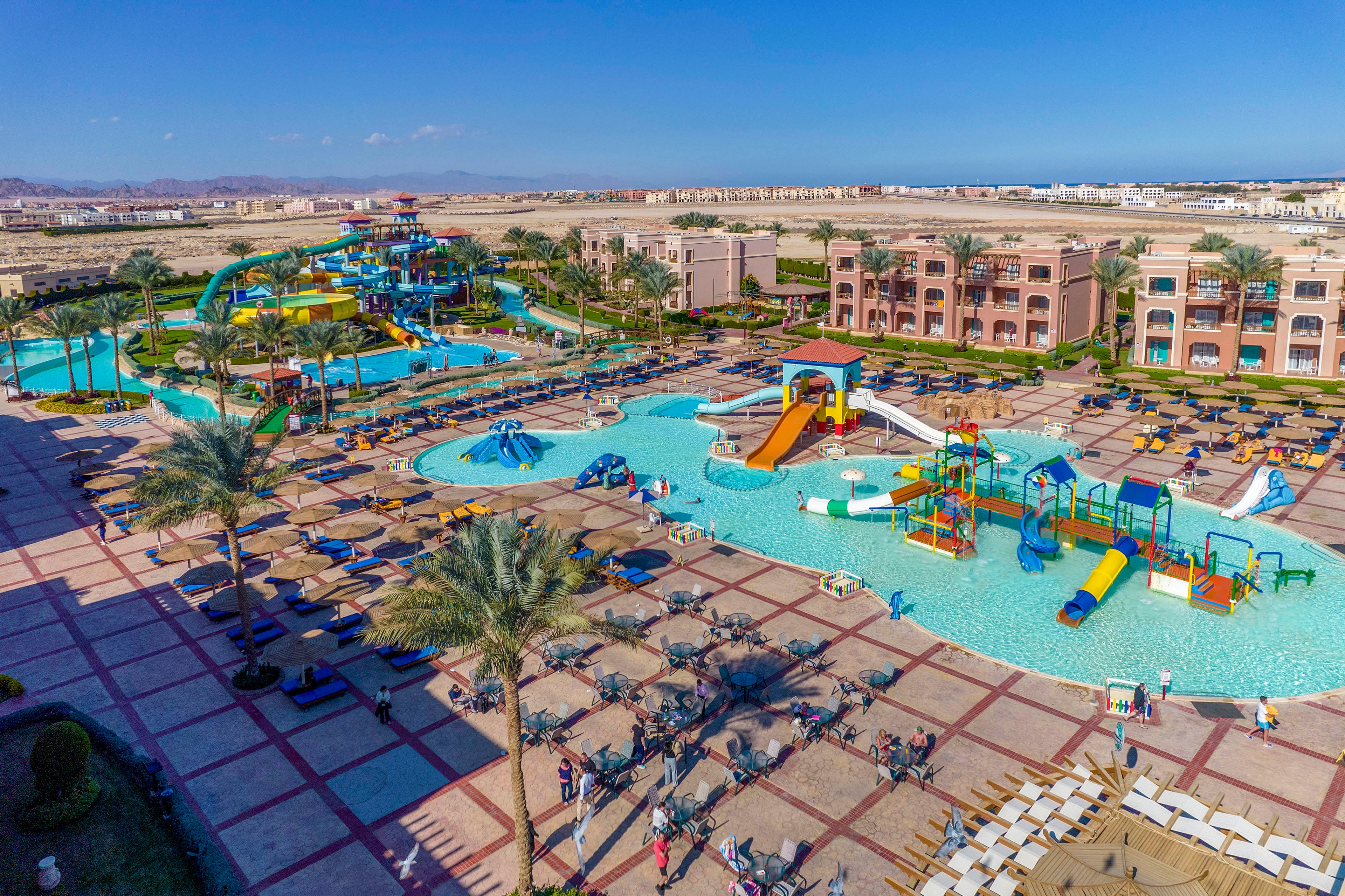 An expansive water park resort featuring multiple pools, colorful water slides, and play areas. The setting includes sun loungers, umbrellas, and palm trees, creating a lively and family-friendly atmosphere. The surrounding architecture showcases modern resort buildings under a clear blue sky.
