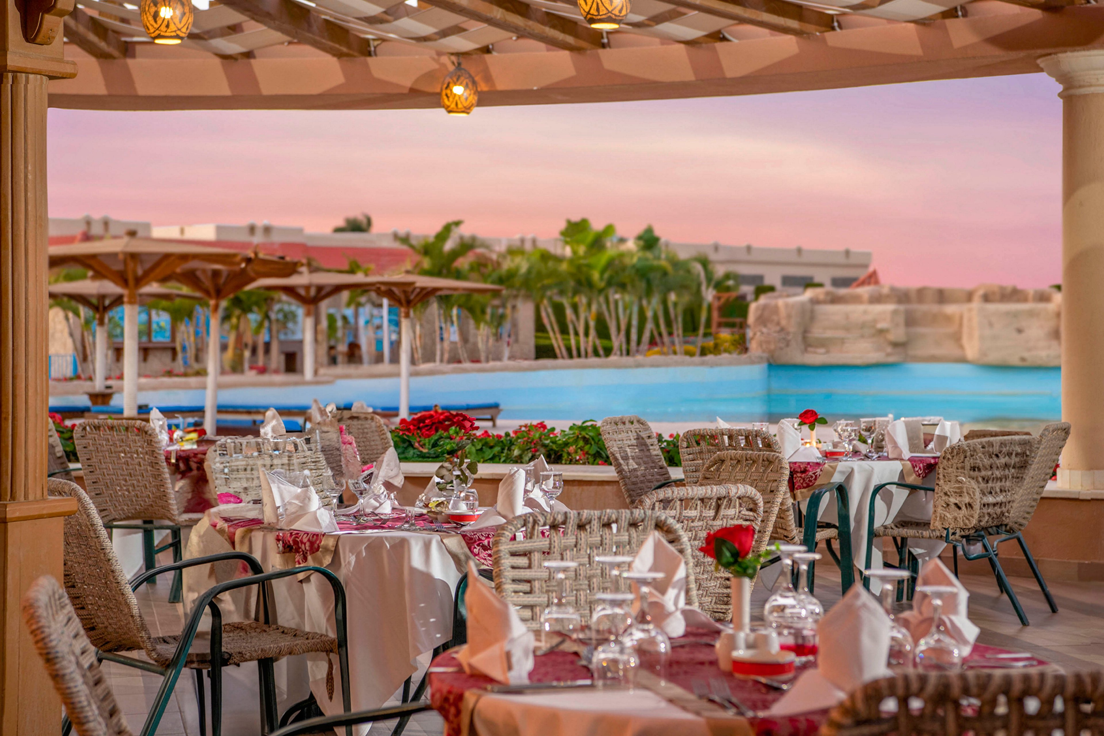 An elegant outdoor dining area set with tables and chairs overlooks a serene pool under a pink-hued sunset sky. The tables are adorned with white tablecloths, red accents, and neatly arranged glassware and napkins. The setting features lush greenery and a resort-like ambiance, creating a relaxing and luxurious atmosphere.