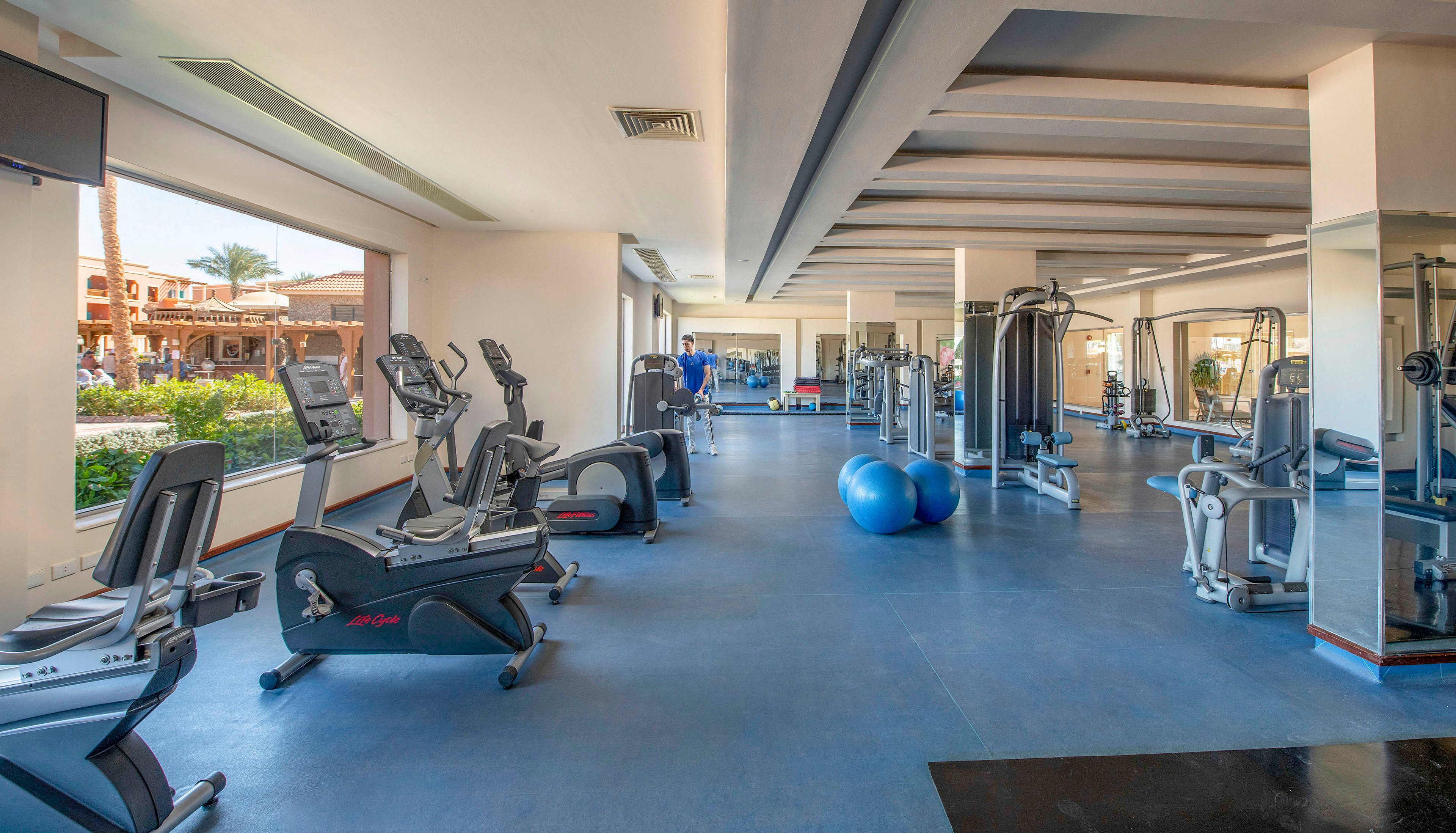 A spacious gym featuring various exercise machines, including treadmills and stationary bikes, with a woman using one of the machines. The room is well-lit with large windows offering views of an outdoor area. Blue exercise balls and other fitness equipment are visible, creating a clean and organized workout environment.