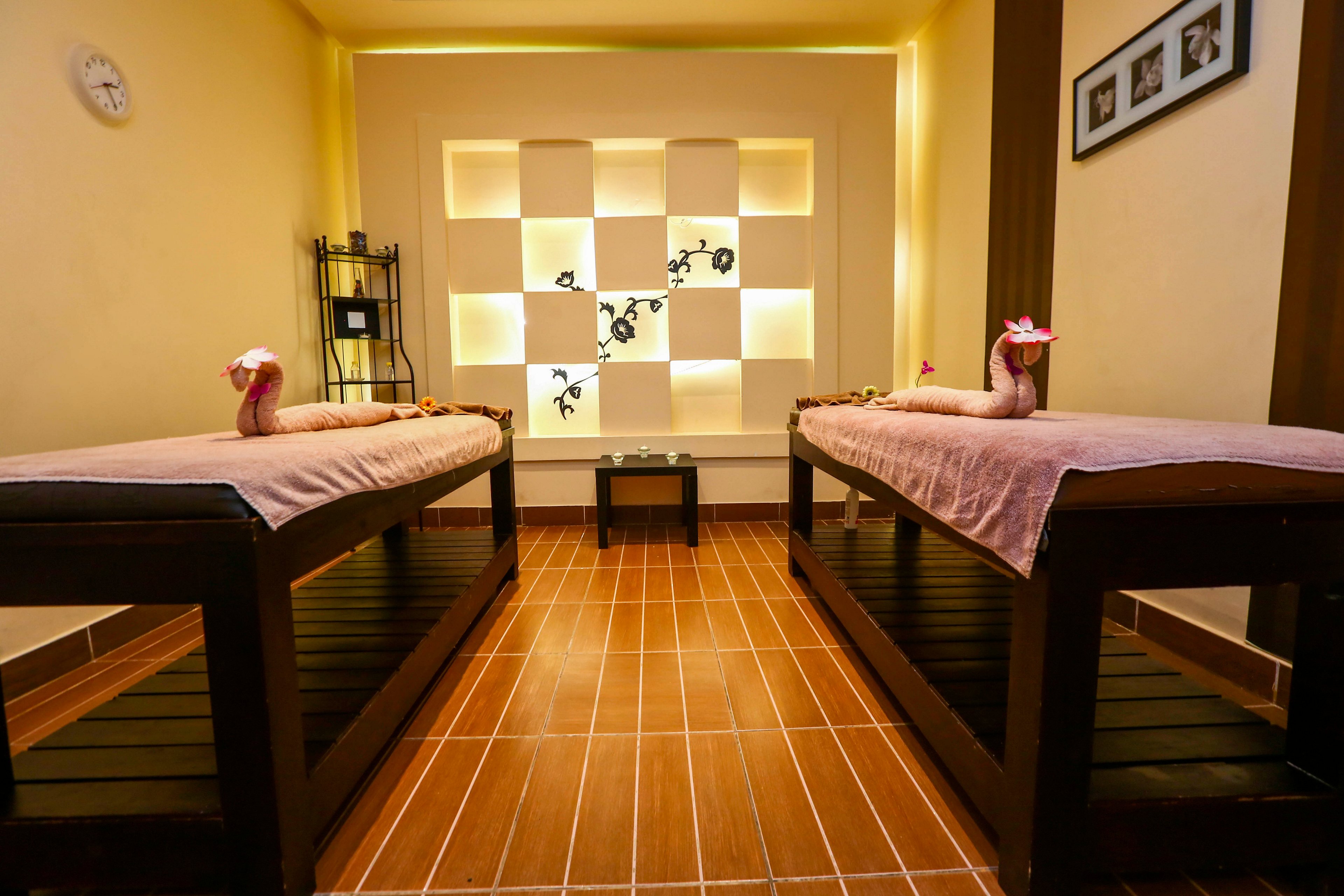 A serene spa room featuring two neatly arranged massage tables with pink towels and decorative flowers. The warm lighting and wooden flooring create a calming ambiance. A modern wall shelf with decorative elements adds to the aesthetic appeal.