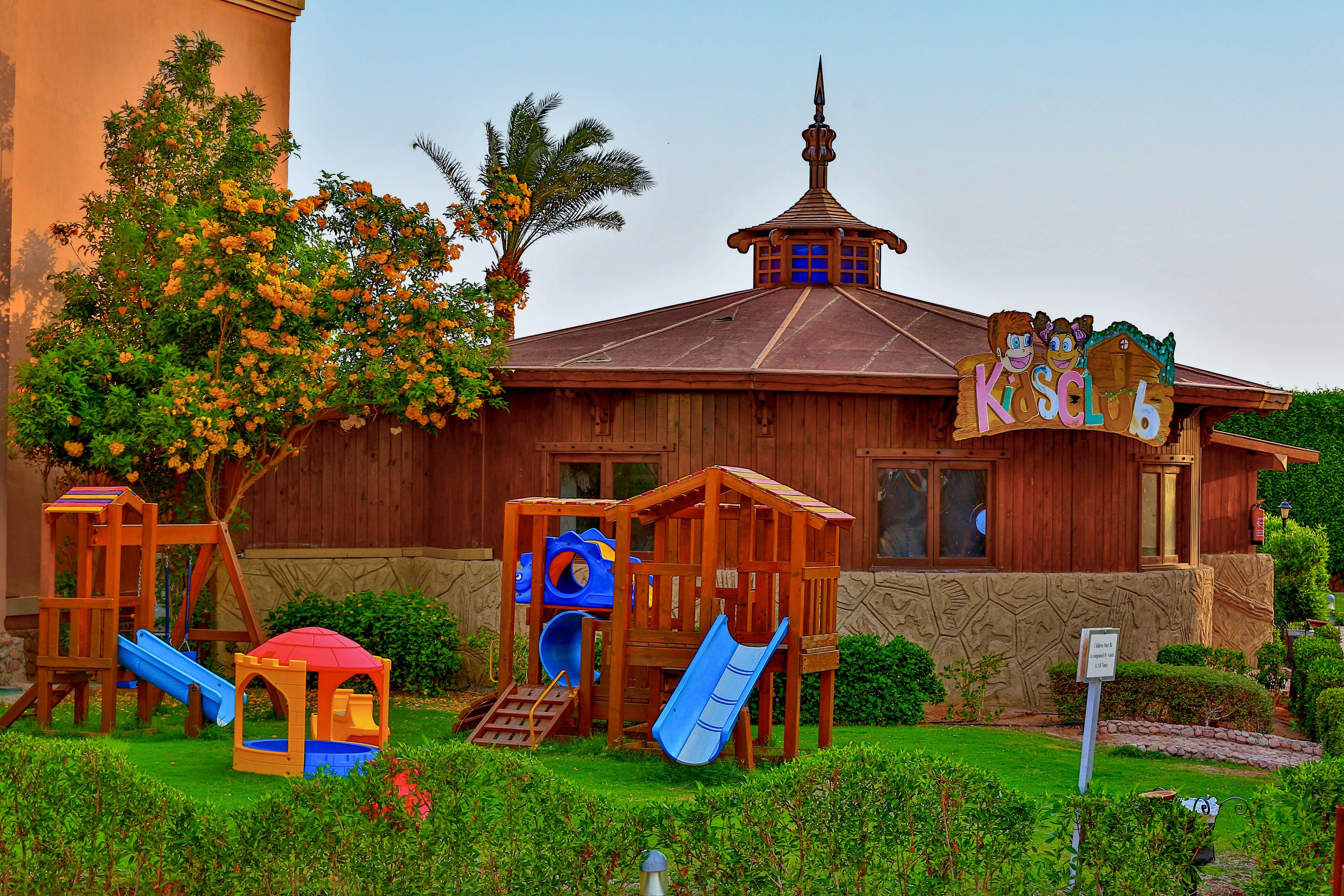 A vibrant children's play area featuring wooden play structures, slides, and a small playhouse. The setting is surrounded by lush greenery and a wooden building with a sign reading 'Kids Club'. The warm lighting enhances the inviting atmosphere.