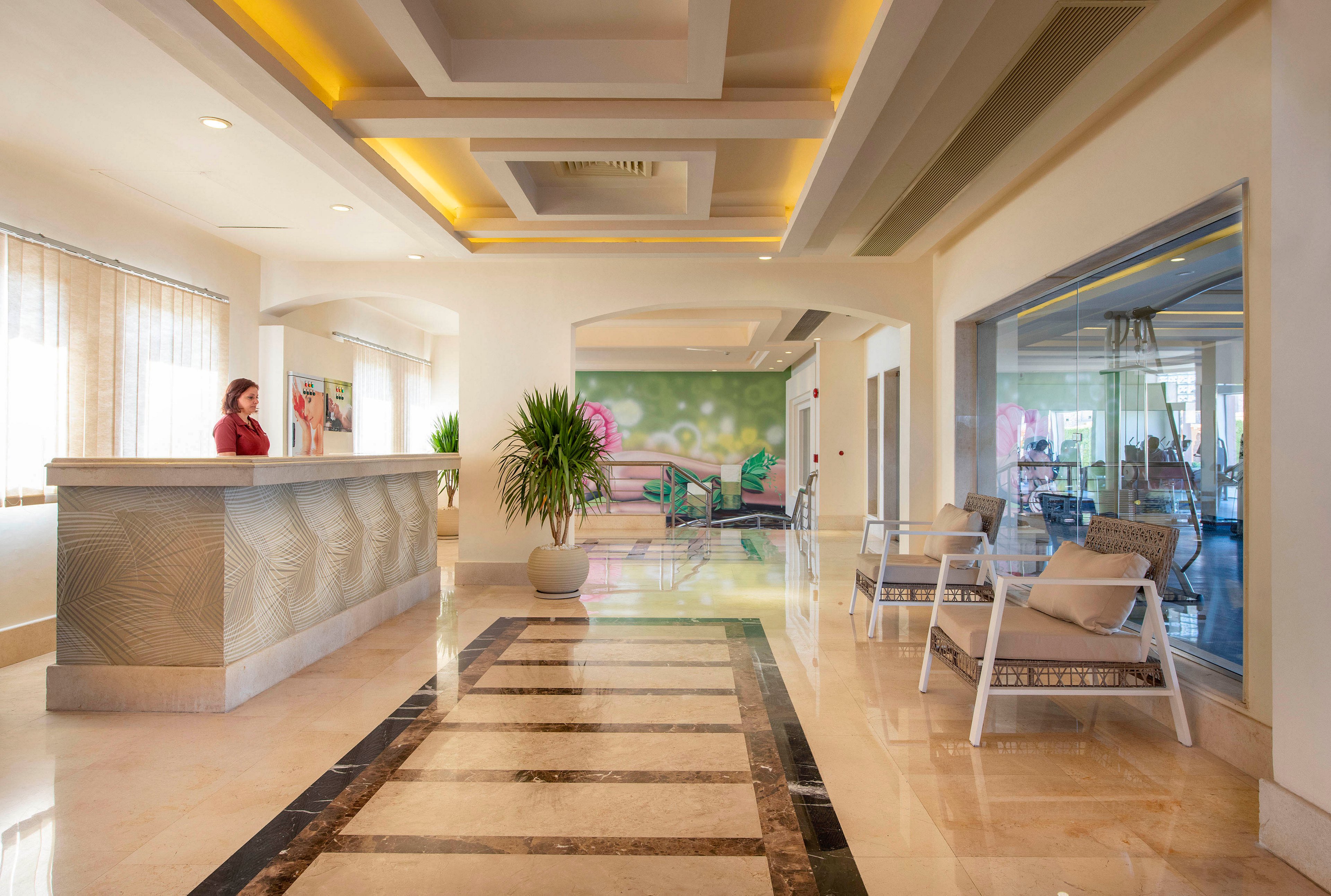 A spacious and well-lit hotel lobby featuring a modern reception desk with a staff member standing behind it. The area includes comfortable seating, decorative plants, and a polished marble floor. The ceiling design and warm lighting create an inviting atmosphere.