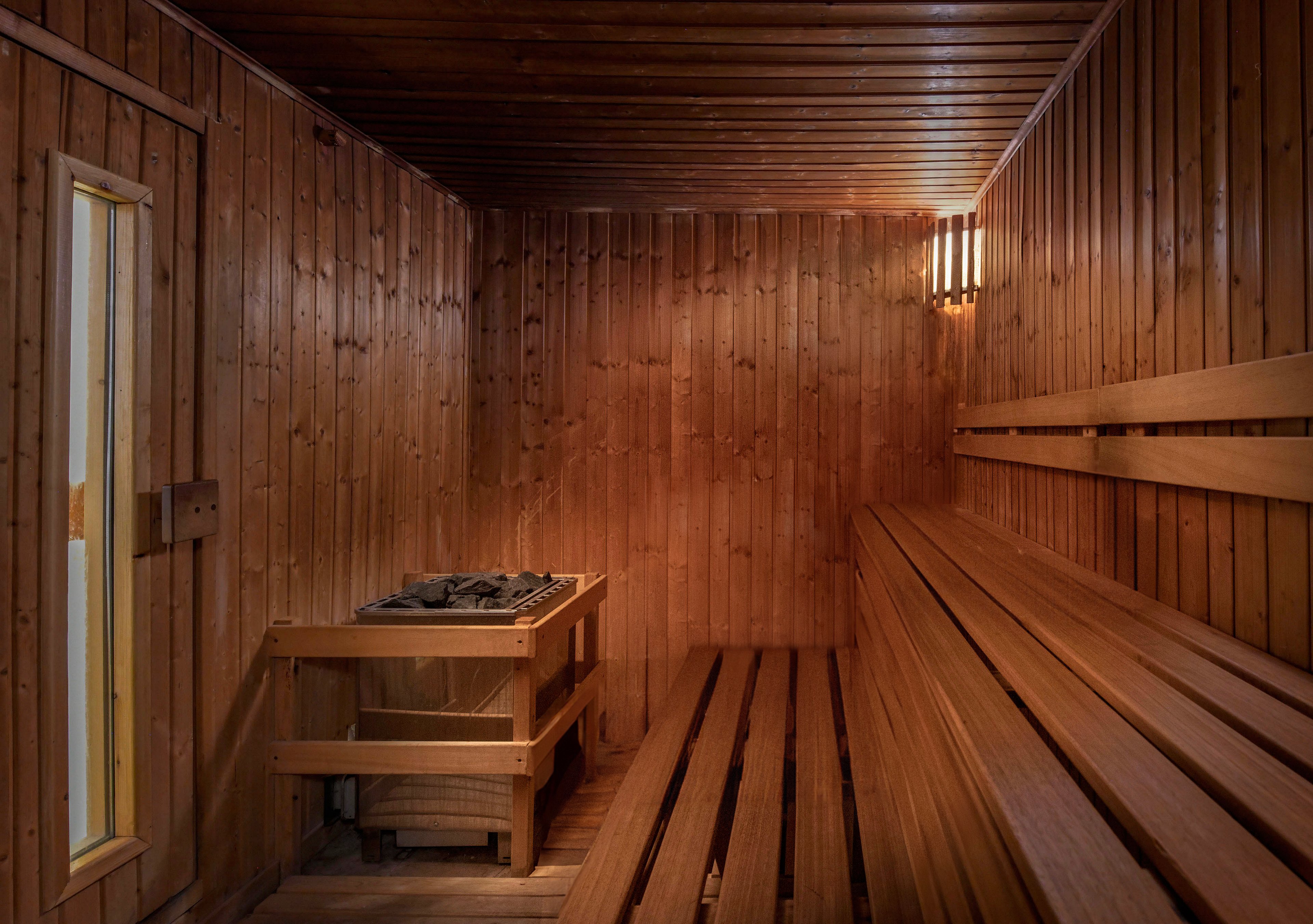 A woman in a teal bikini sits on a wooden bench inside a traditional sauna room. The setting features wooden walls, a heater with stones, and warm lighting, creating a cozy and tranquil atmosphere. The scene emphasizes relaxation and wellness in a spa-like environment.