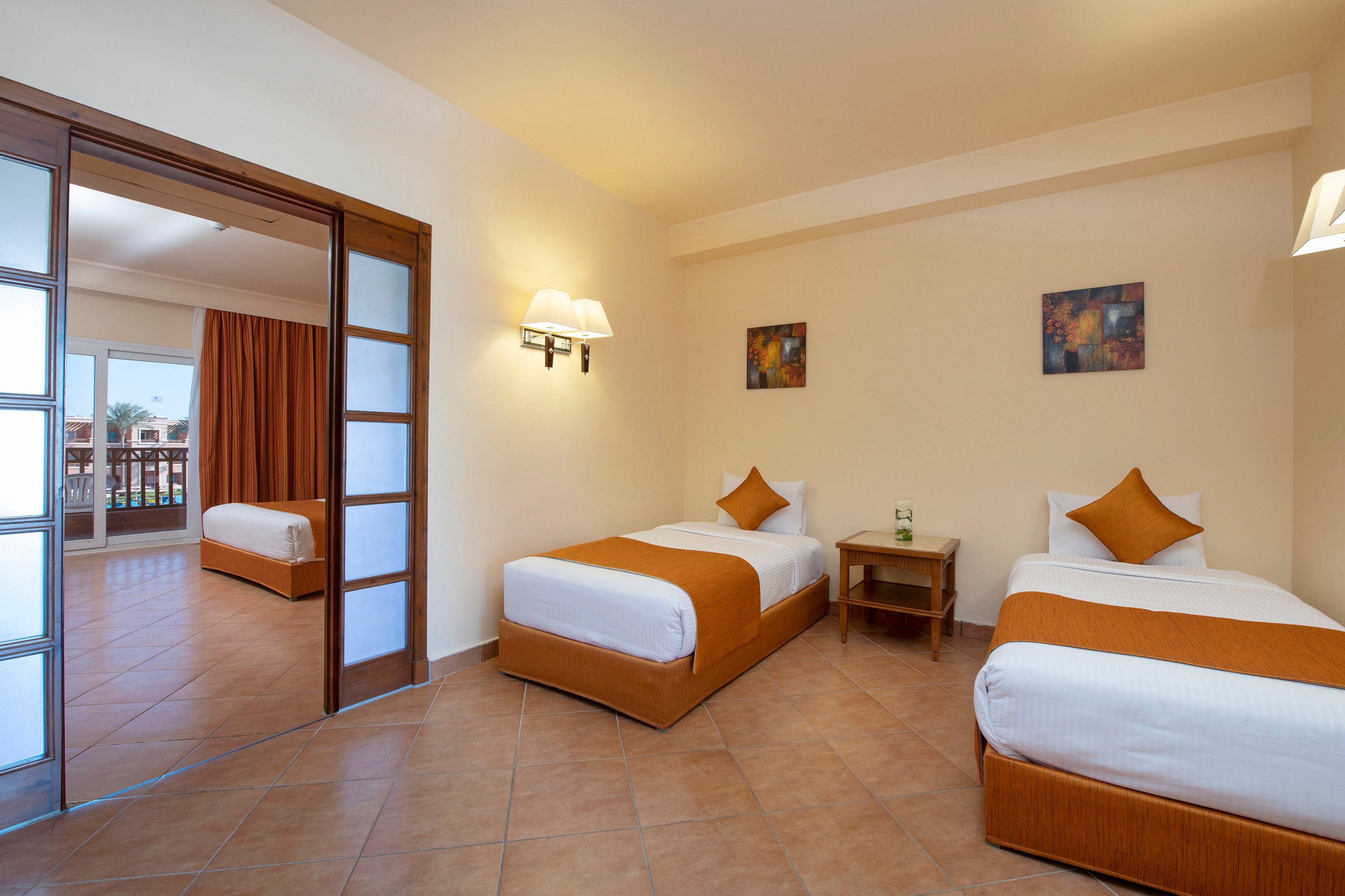 A bright and spacious hotel room featuring two twin beds with orange accents and a wooden side table. The room has tiled flooring, warm lighting, and wall art for a cozy ambiance. A sliding door opens to an adjoining room with a single bed and a balcony view.