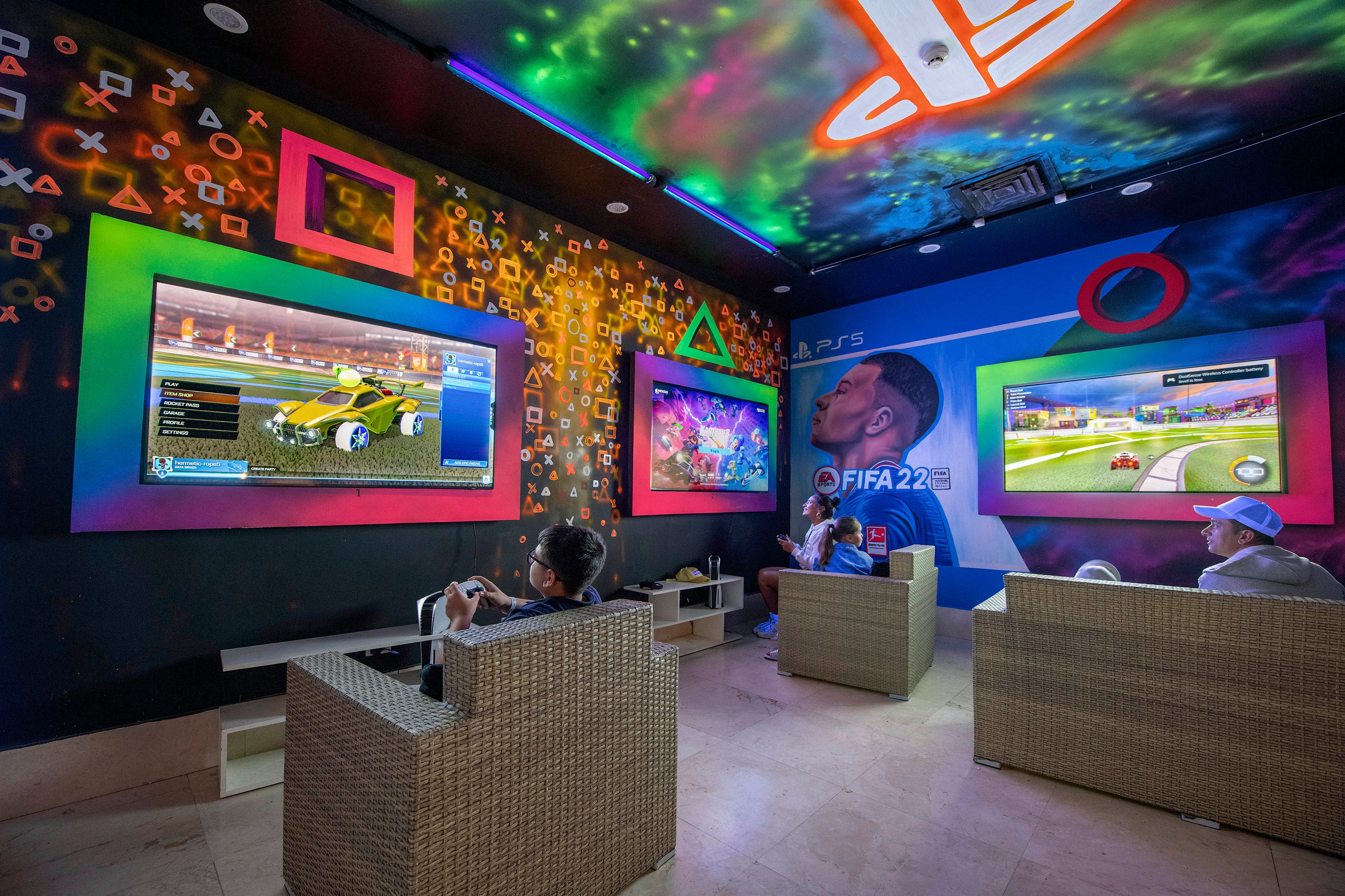 A modern gaming lounge featuring multiple screens displaying video games, including FIFA 22. The setting includes colorful wall art with gaming symbols and comfortable seating. The atmosphere is lively and immersive, with players engaged in gameplay. Visible text includes 'FIFA 22' and PlayStation symbols.