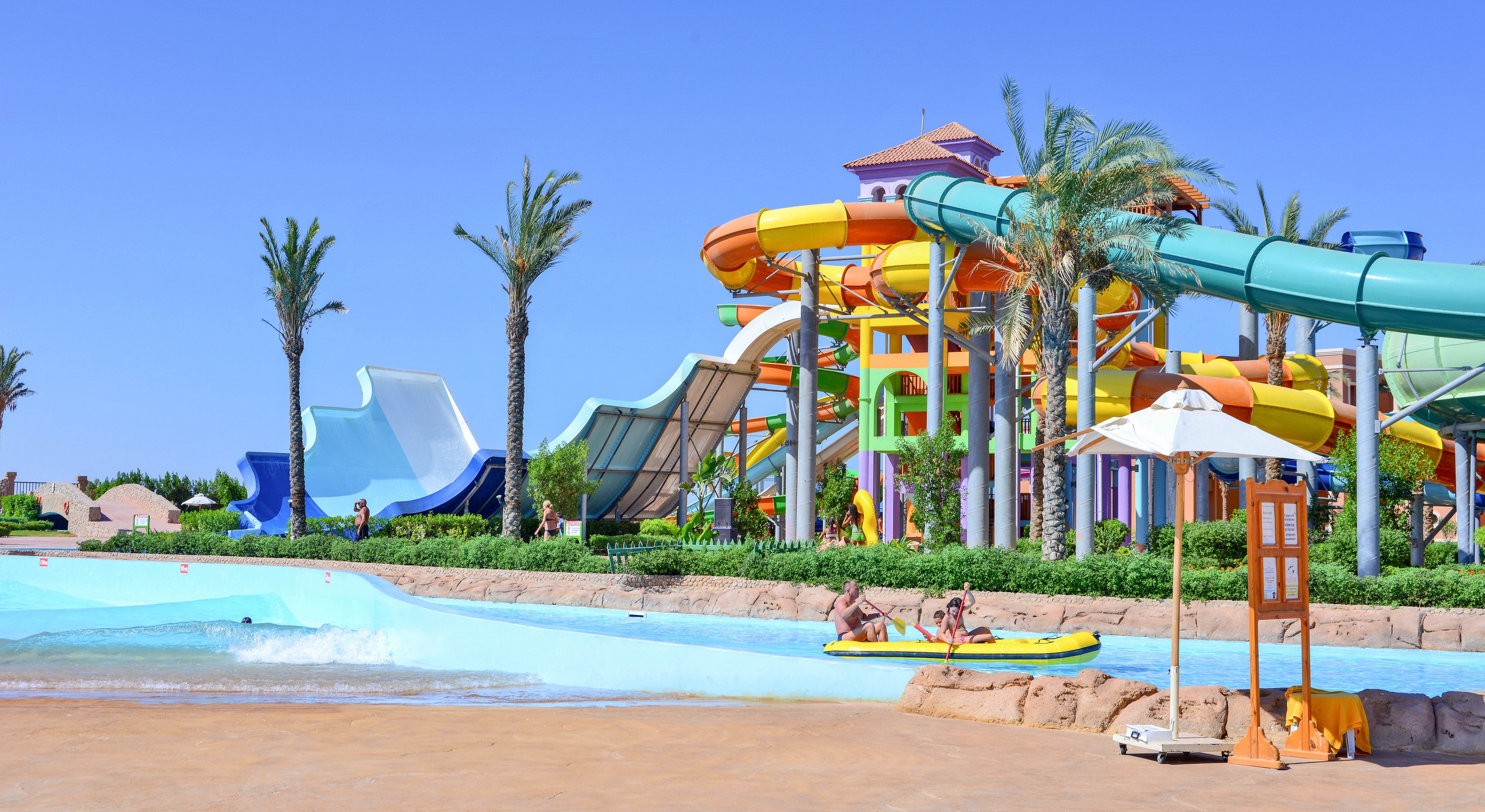 A vibrant water park featuring colorful slides, a wave pool, and palm trees under a clear blue sky. Visitors enjoy the attractions, including a yellow inflatable raft in the pool. The setting is lively and family-friendly, with a mix of fun and relaxation.