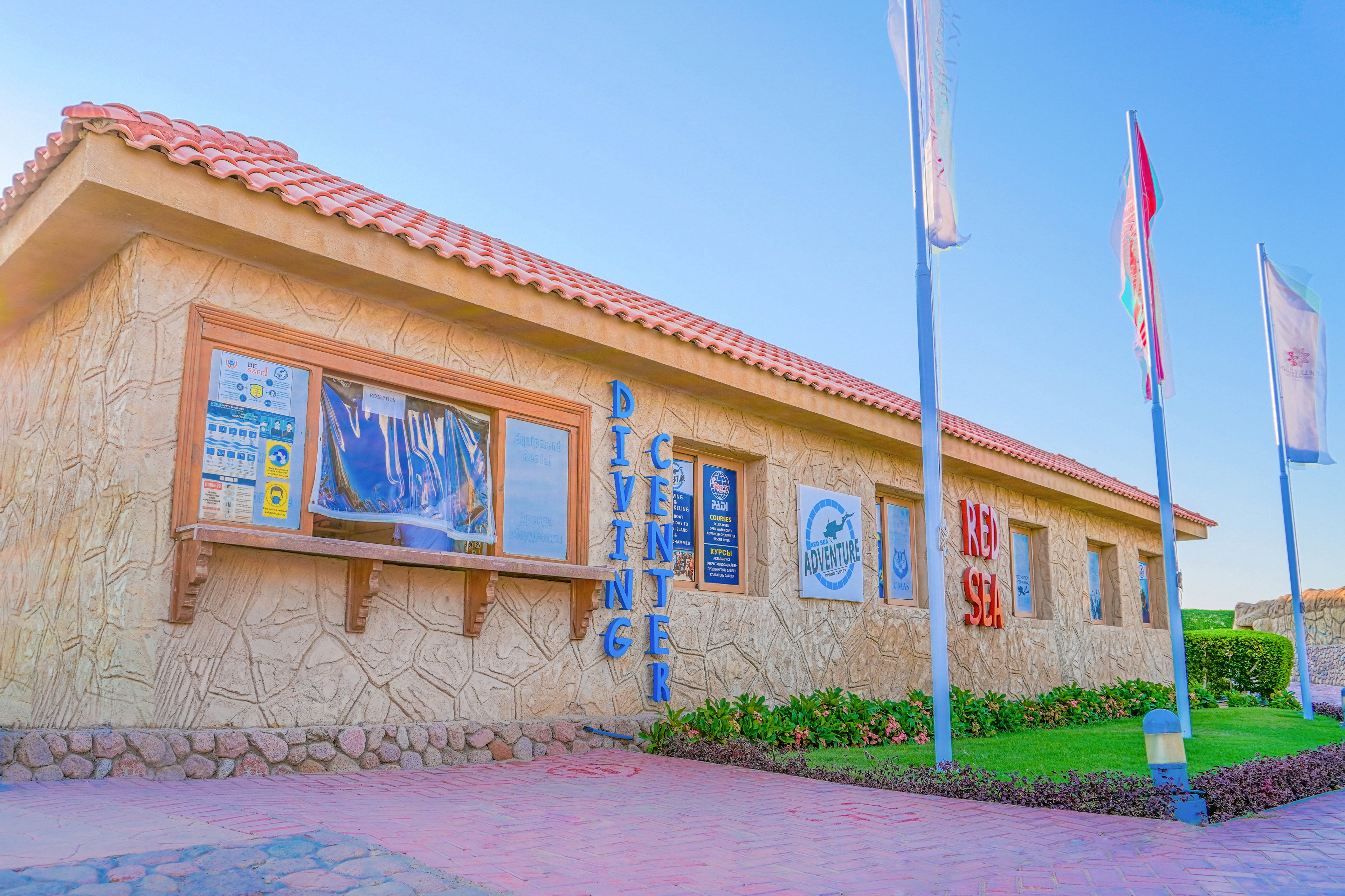 A stone-textured diving center building with prominent signage reading 'Diving Center' and 'Red Sea'. The setting features a clear blue sky, flags, and a well-maintained garden. Informational posters and logos are visible on the building's exterior.