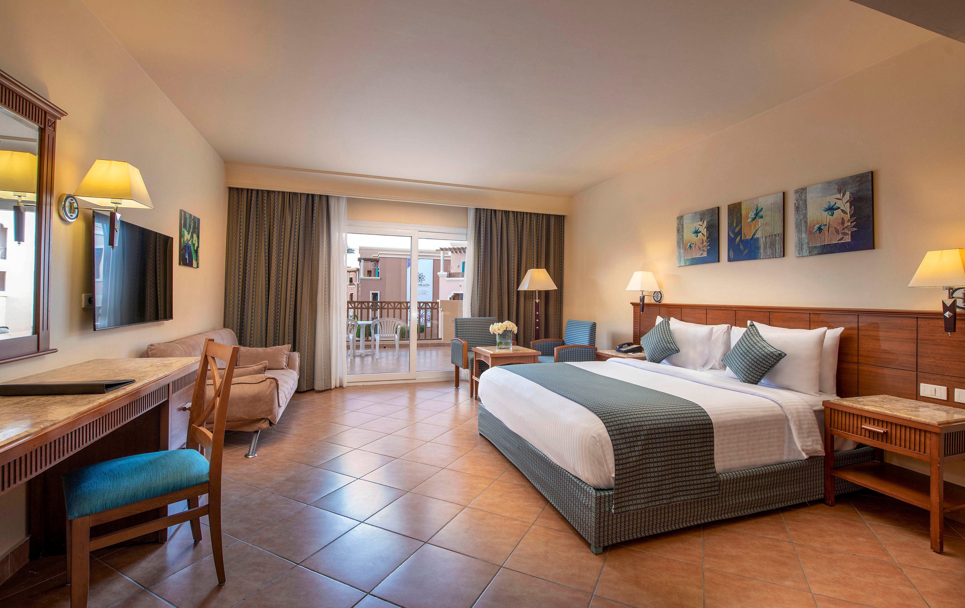 A well-lit hotel room featuring a large bed with white and teal bedding, a seating area, and a desk. The room opens to a balcony with a view of neighboring buildings. Warm lighting and tiled flooring create a cozy and inviting atmosphere.