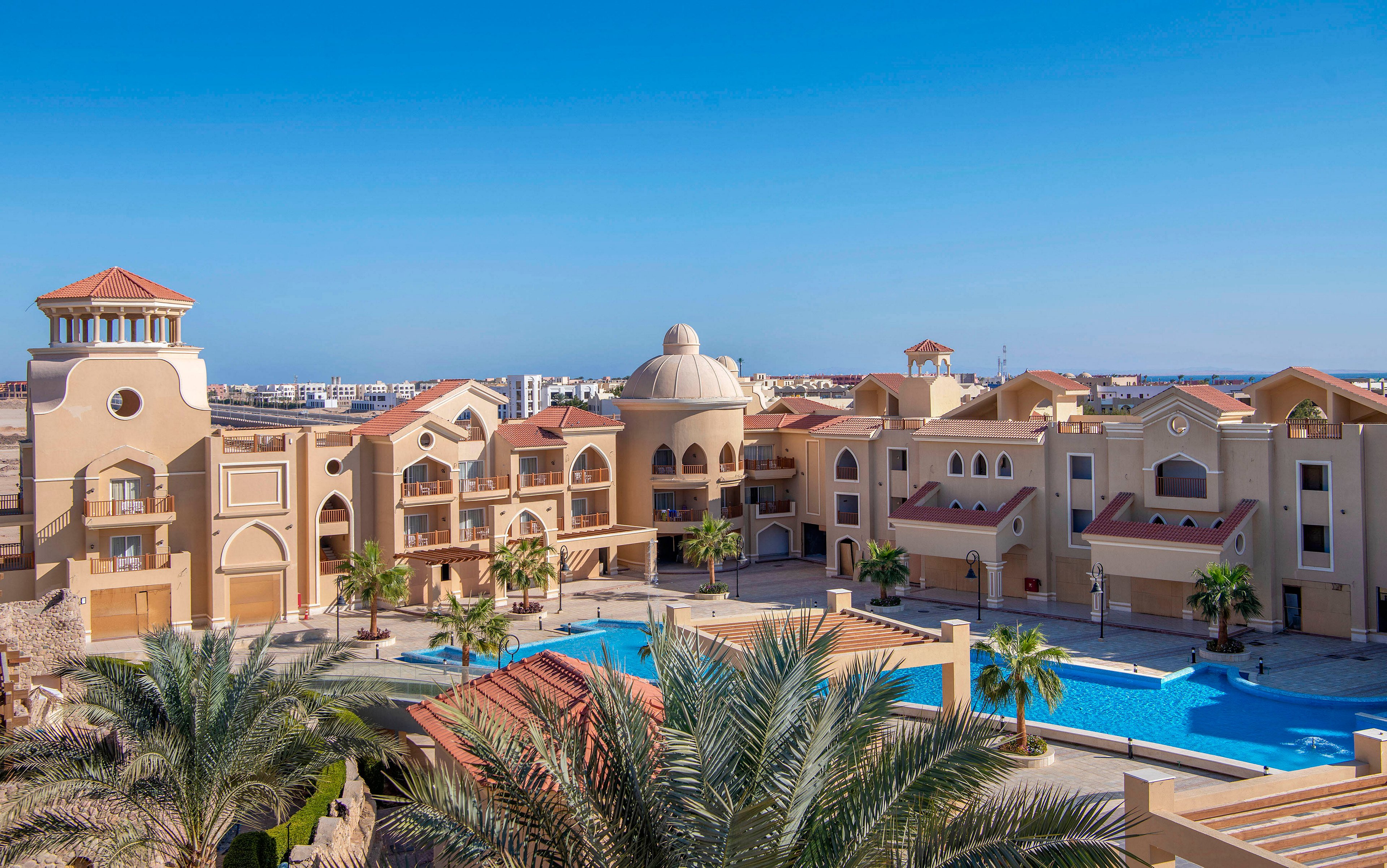 A luxurious resort featuring Mediterranean-style architecture with beige and terracotta tones. The setting includes a large swimming pool surrounded by palm trees and elegant buildings under a clear blue sky. The tranquil atmosphere suggests a relaxing vacation destination.