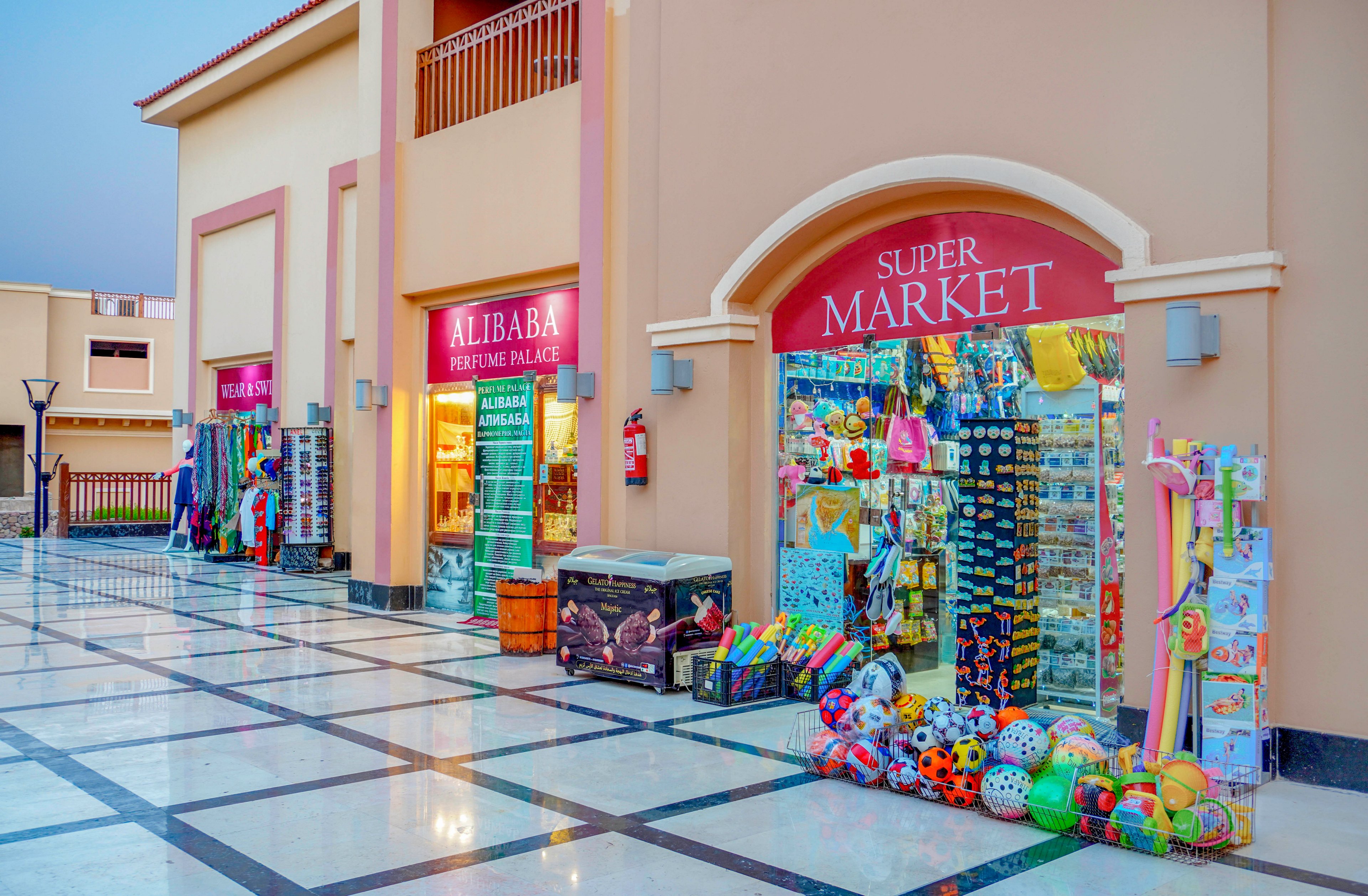 A vibrant supermarket storefront featuring a variety of colorful toys and goods displayed outside. The setting is a tiled walkway with a warm, pastel-colored building facade. Visible signage includes 'Super Market' and 'Alibaba Perfume Palace,' adding to the lively atmosphere.