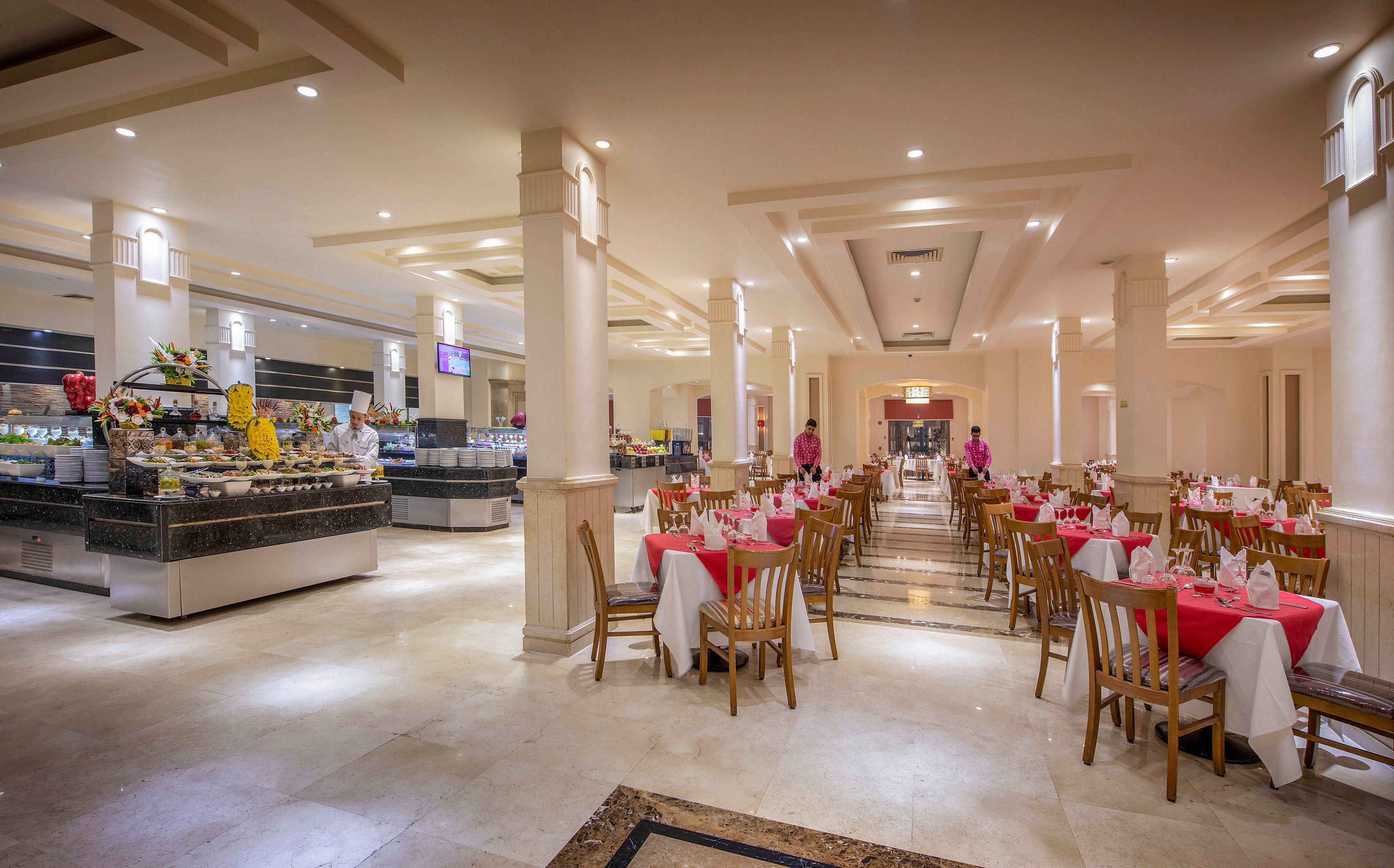 A spacious and well-lit restaurant dining area featuring neatly arranged tables with red and white tablecloths. The buffet section is visible on the left, showcasing a variety of food items and decorative displays. The setting exudes a warm and inviting atmosphere, ideal for formal dining or gatherings.