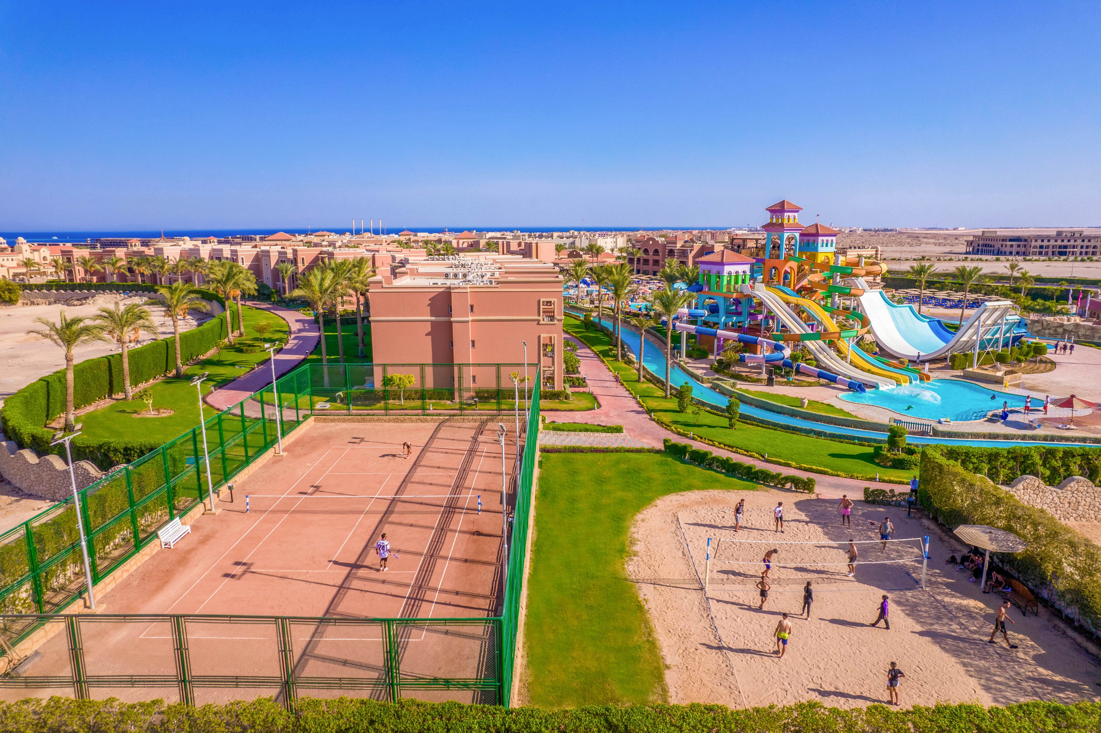 A vibrant resort featuring tennis courts, a sandy volleyball area, and a colorful water park with slides and pools. The setting is sunny and spacious, surrounded by palm trees and well-maintained landscaping. The scene captures leisure activities and outdoor fun in a tropical environment.