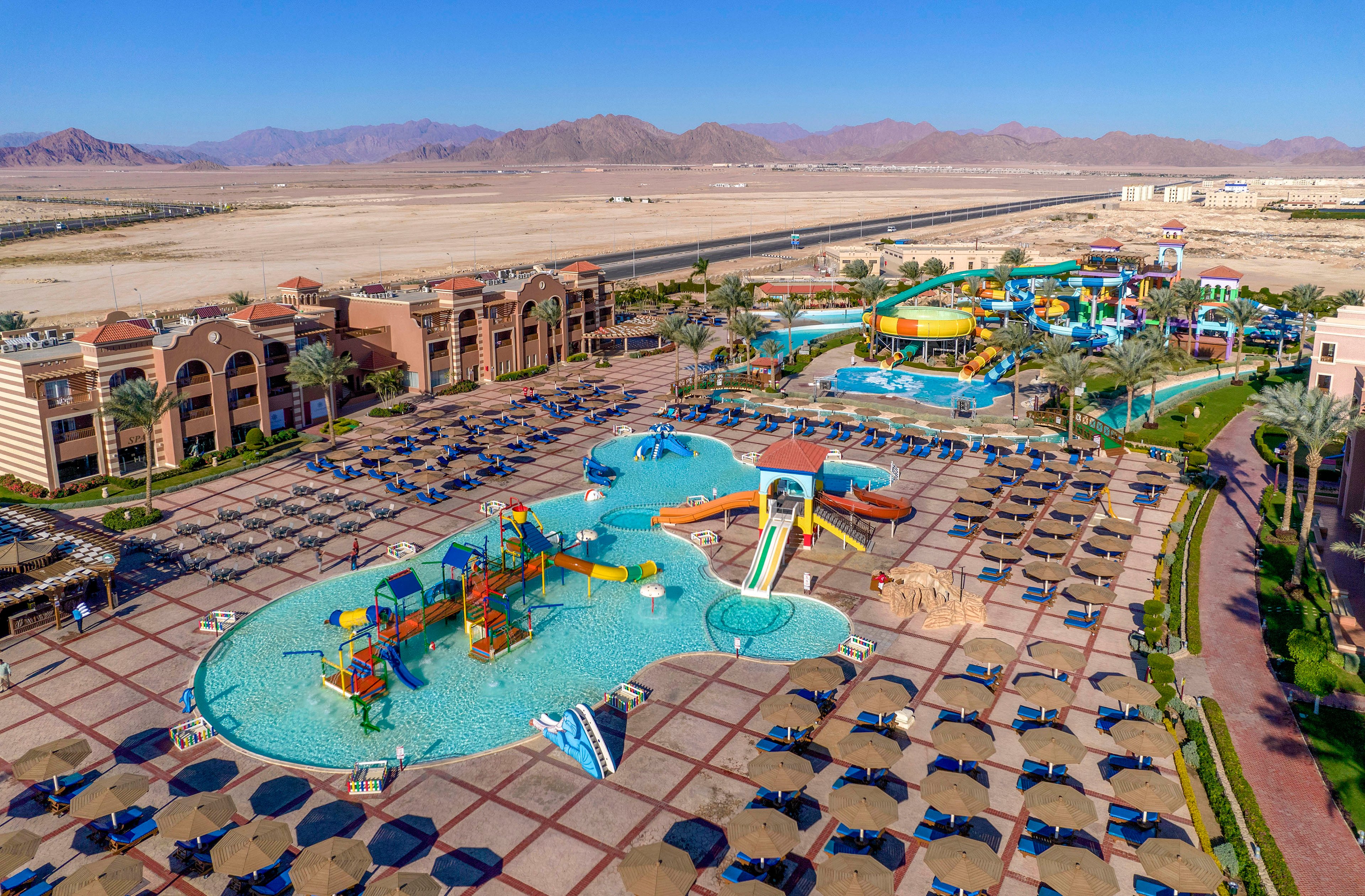 A vibrant aerial view of a resort featuring a large water park with slides, pools, and colorful play structures. The setting includes neatly arranged sun loungers and umbrellas, surrounded by palm trees and desert mountains in the background. The clear blue sky enhances the lively and inviting atmosphere.