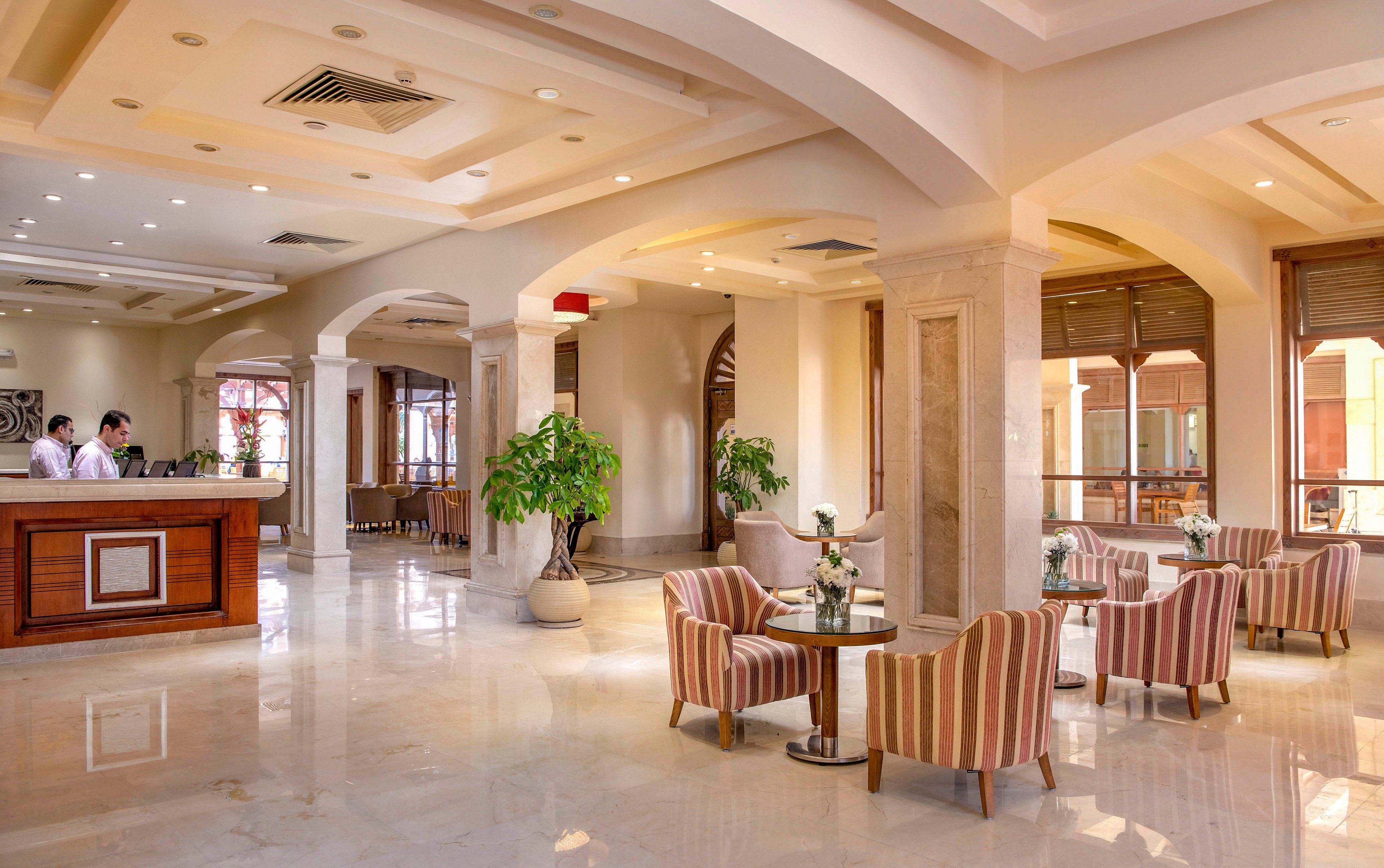 A spacious and well-lit hotel lobby featuring a reception desk and a cozy seating area with striped armchairs and small tables. The interior is adorned with potted plants, marble flooring, and arched windows allowing natural light. The warm beige tones and soft lighting create a welcoming and luxurious ambiance.
