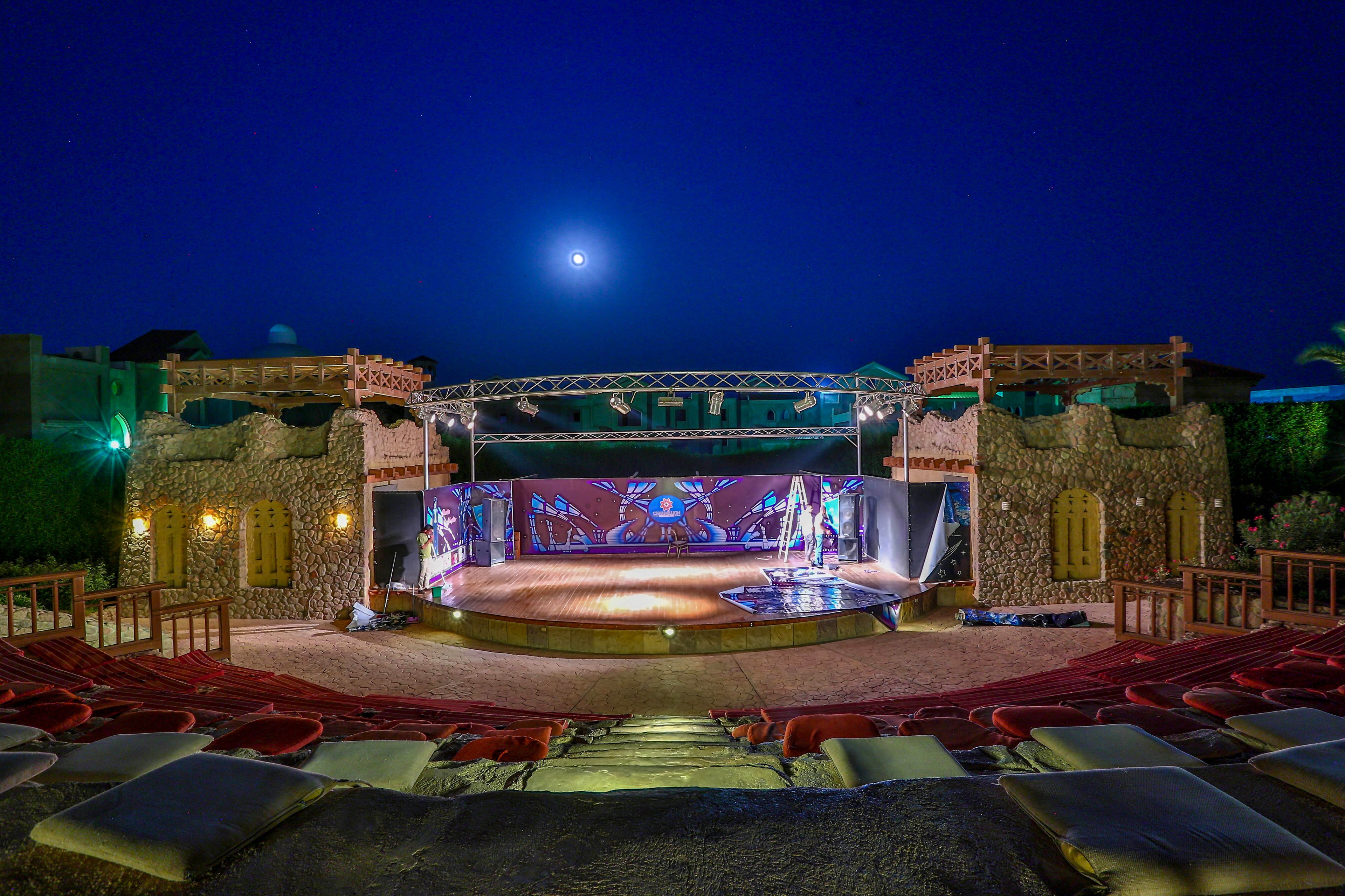 A beautifully lit outdoor amphitheater with a stage set against a backdrop of traditional architectural elements. The scene is illuminated by soft lighting and a full moon in the night sky. The seating area features cushioned benches, creating a cozy and inviting atmosphere for performances.