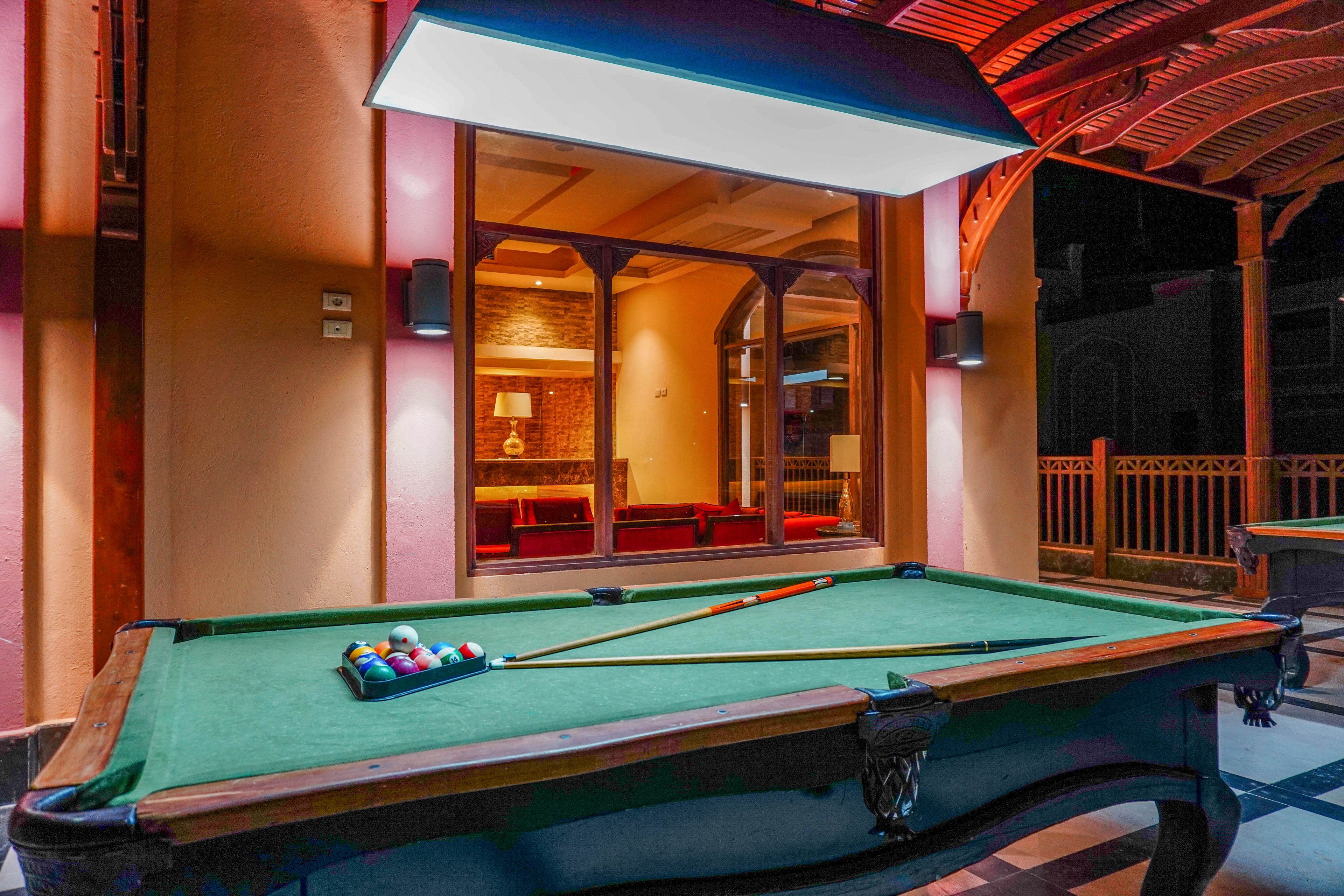 A well-lit outdoor billiards table with a green felt surface is set under a canopy in a warm, inviting space. The table is equipped with a cue stick and a rack of billiard balls, ready for play. The background features a window revealing a cozy indoor seating area with warm lighting and wooden accents.