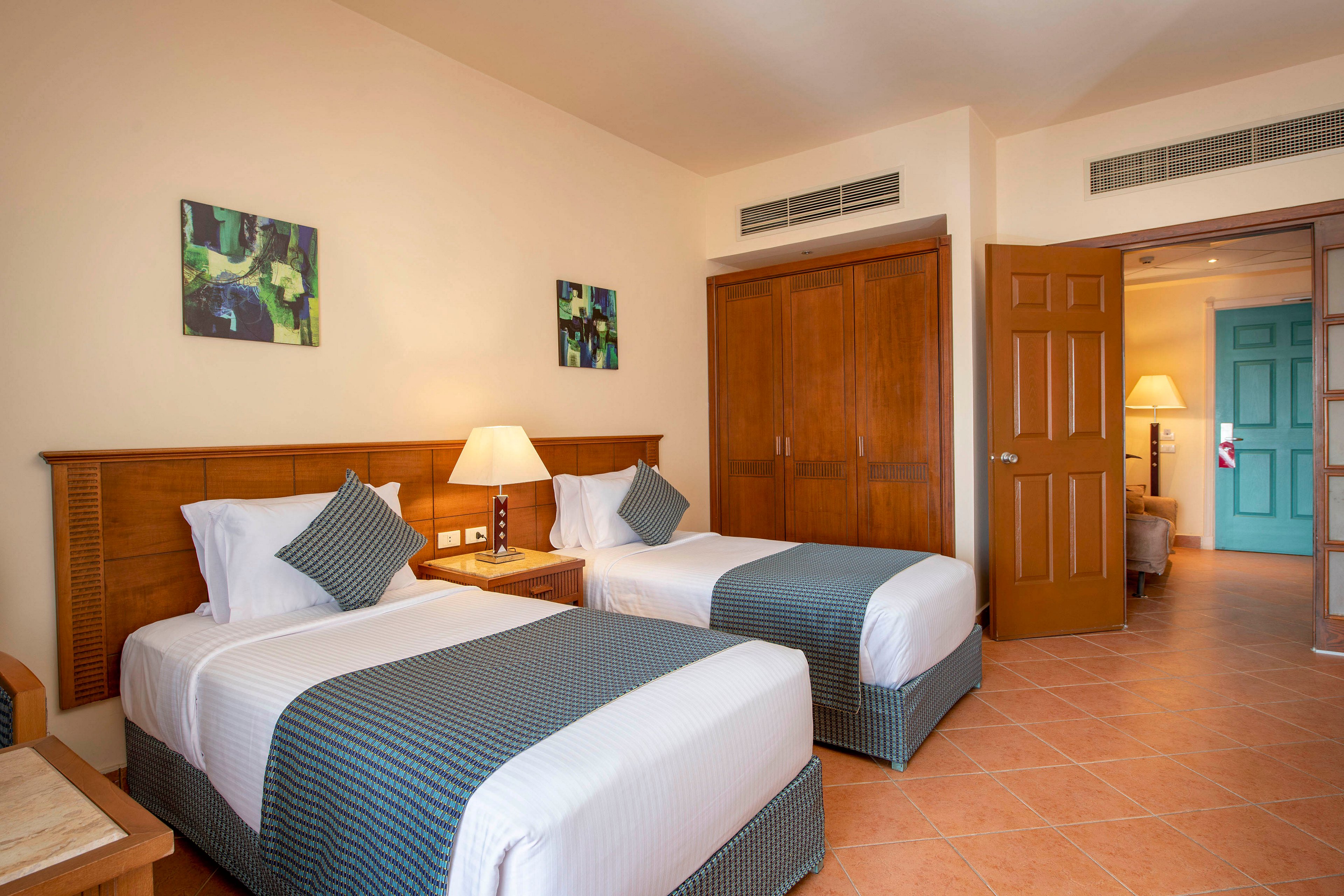 A well-lit hotel room featuring two neatly made twin beds with matching blue and white bedding. The room includes wooden furniture, a bedside lamp, and wall art for a warm and inviting ambiance. The open door reveals a glimpse of an adjoining area with additional lighting and decor.