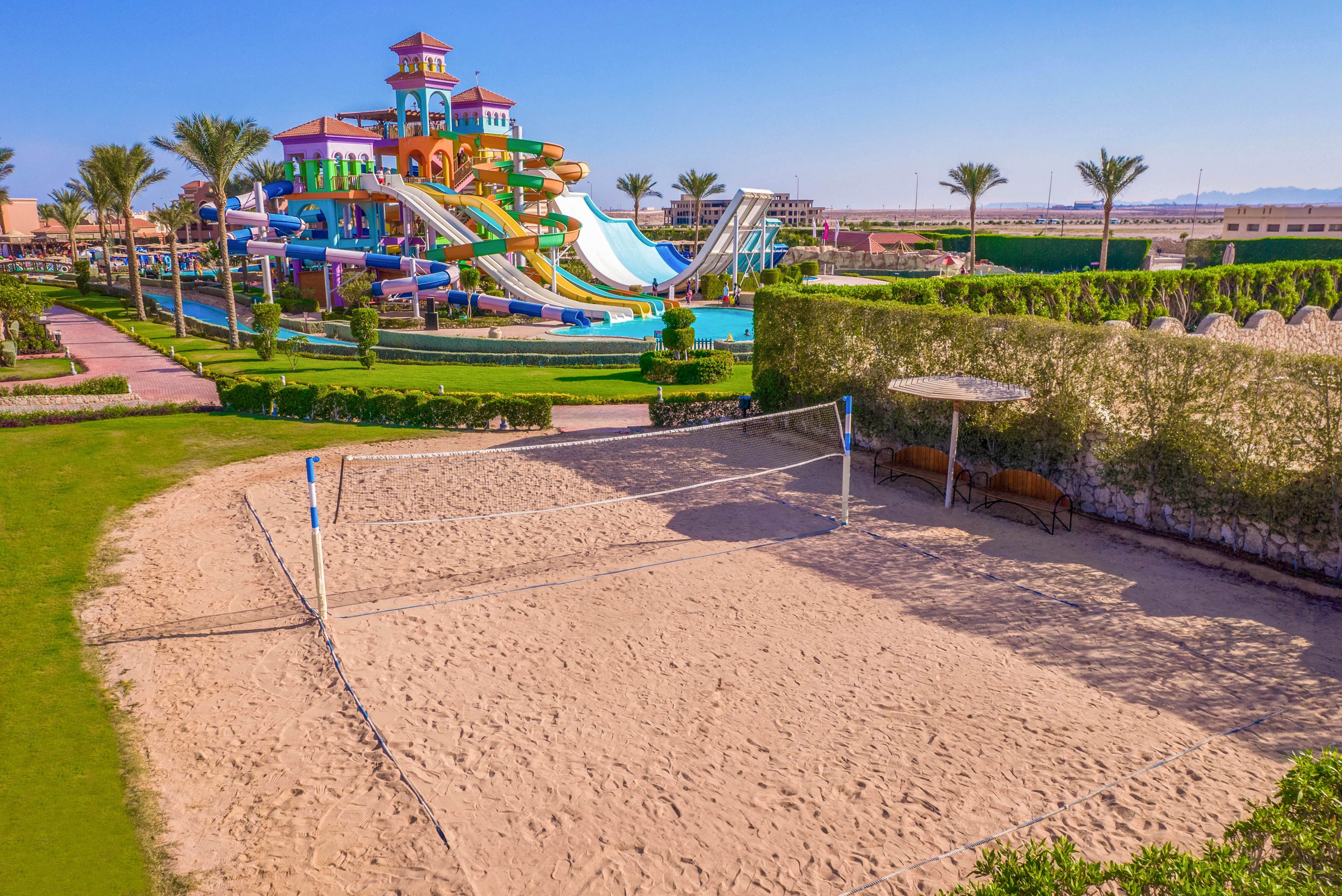 A group of people playing beach volleyball on a sandy court near a vibrant water park with colorful slides and towers. The setting is outdoors on a sunny day, surrounded by palm trees and manicured greenery. The water park features multiple slides and a lively atmosphere, creating a fun and energetic environment.