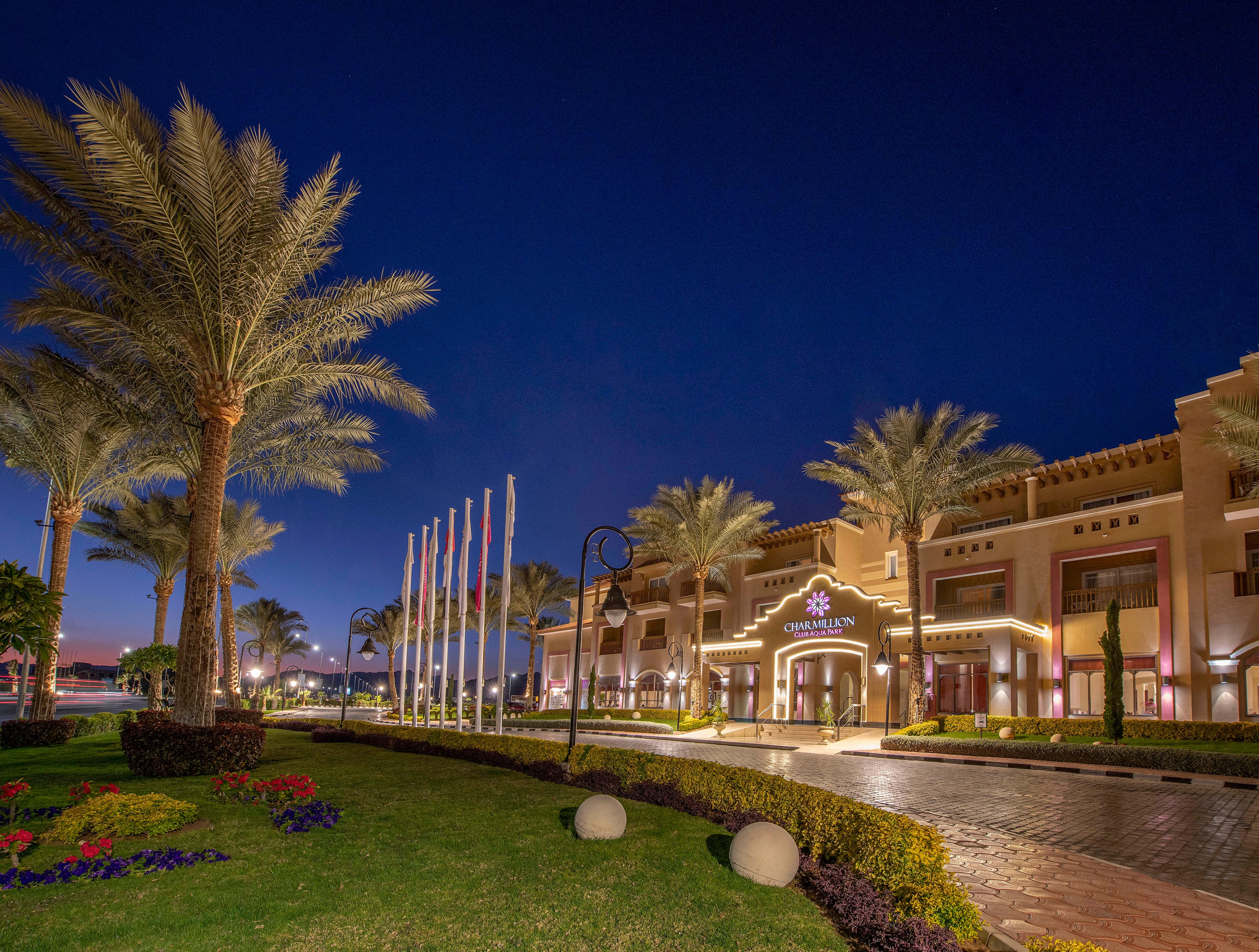 A luxurious hotel entrance illuminated at night, surrounded by well-maintained gardens and tall palm trees. The building features warm lighting and architectural details, with a row of flags displayed prominently. The setting exudes a serene and upscale ambiance.