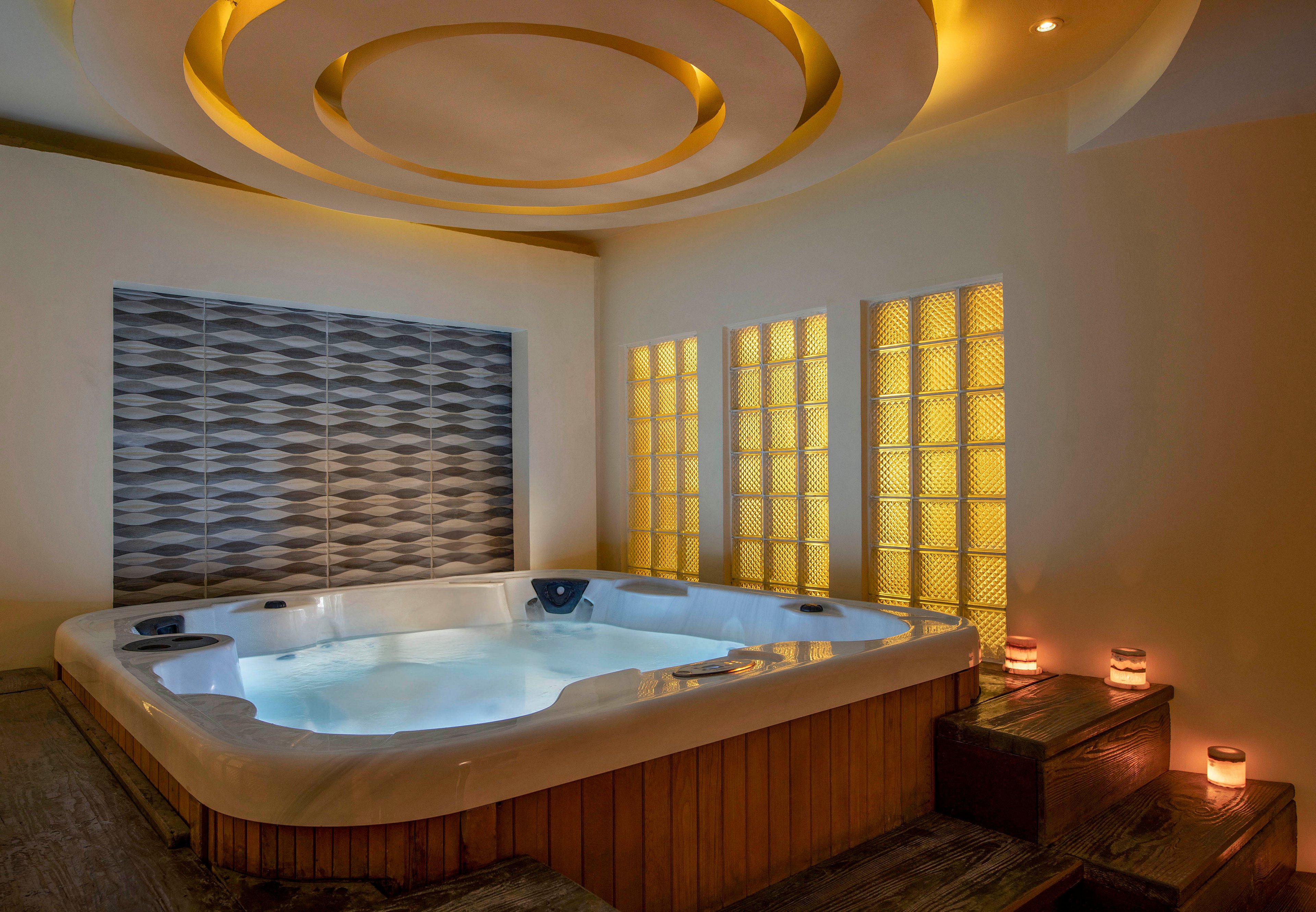 A woman enjoys a relaxing moment in a modern indoor hot tub. The setting features warm lighting, decorative wall panels, and glowing candles, creating a serene ambiance. The circular ceiling design adds a touch of elegance to the spa-like environment.