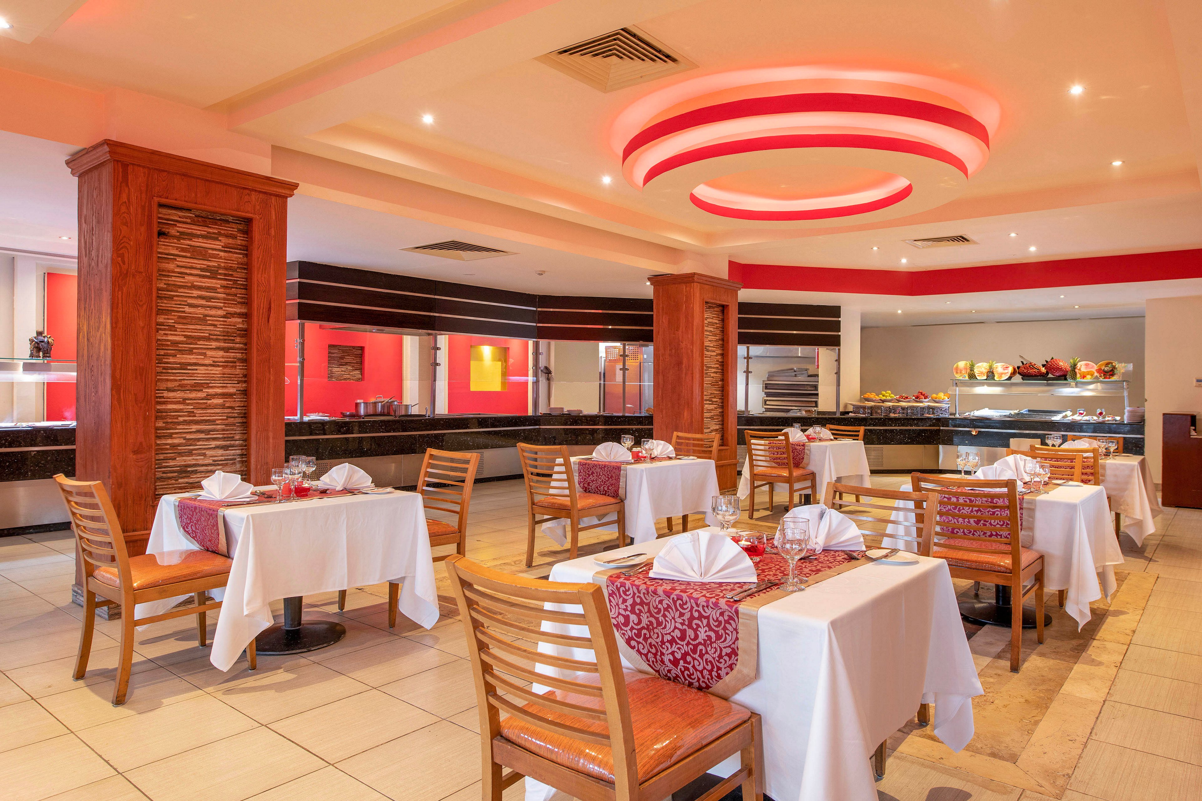 A well-lit restaurant interior featuring neatly arranged dining tables with white tablecloths and red accents. The setting includes wooden chairs, decorative elements, and a buffet area in the background. The ceiling design with circular lighting adds a modern touch to the ambiance.