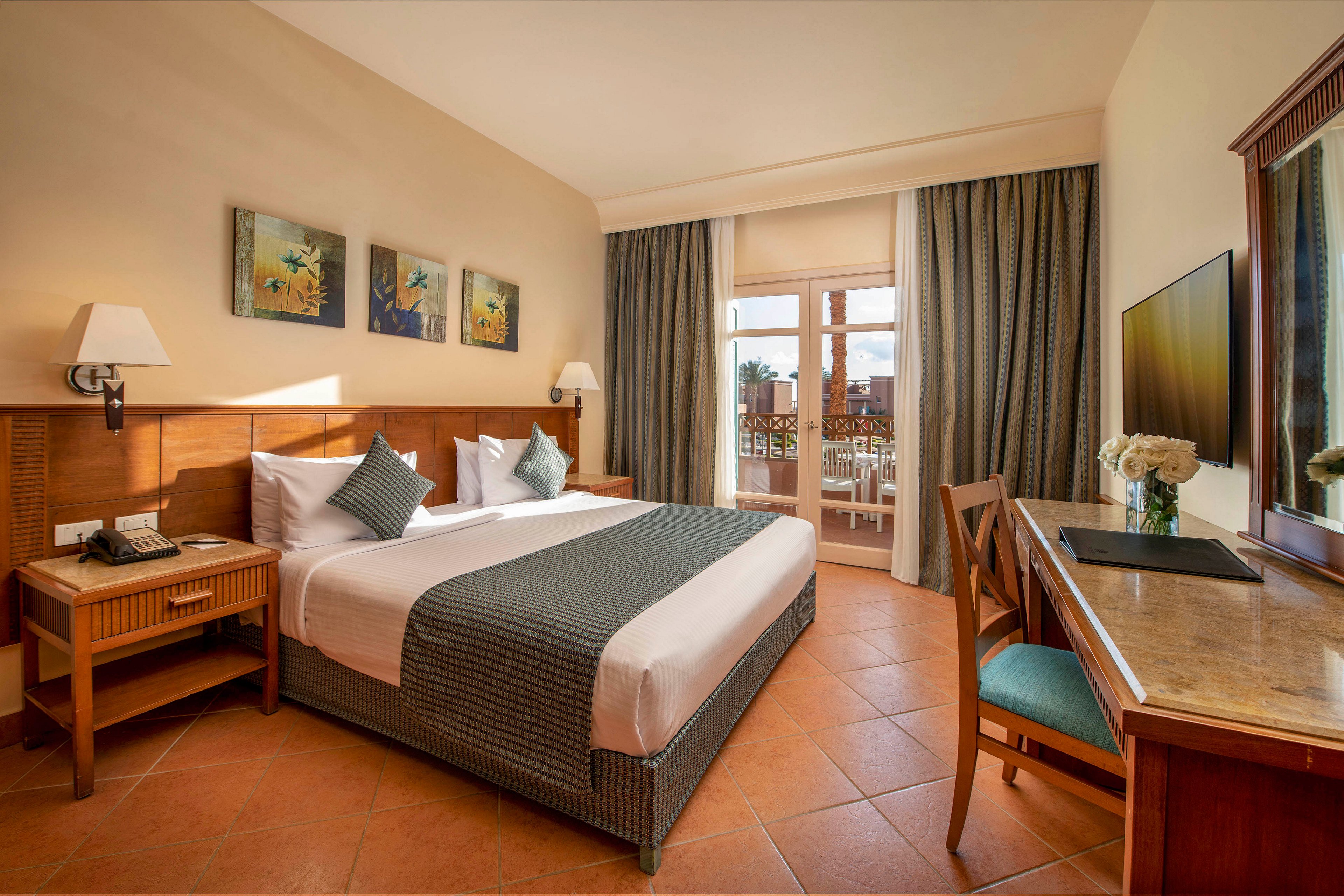 A well-lit hotel room featuring a neatly made double bed with decorative pillows and bedside tables. The room includes a desk with a chair, a flat-screen TV, and a vase of flowers. Large windows open to a balcony with outdoor seating, allowing natural light to fill the space. The warm tones and elegant decor create a welcoming atmosphere.