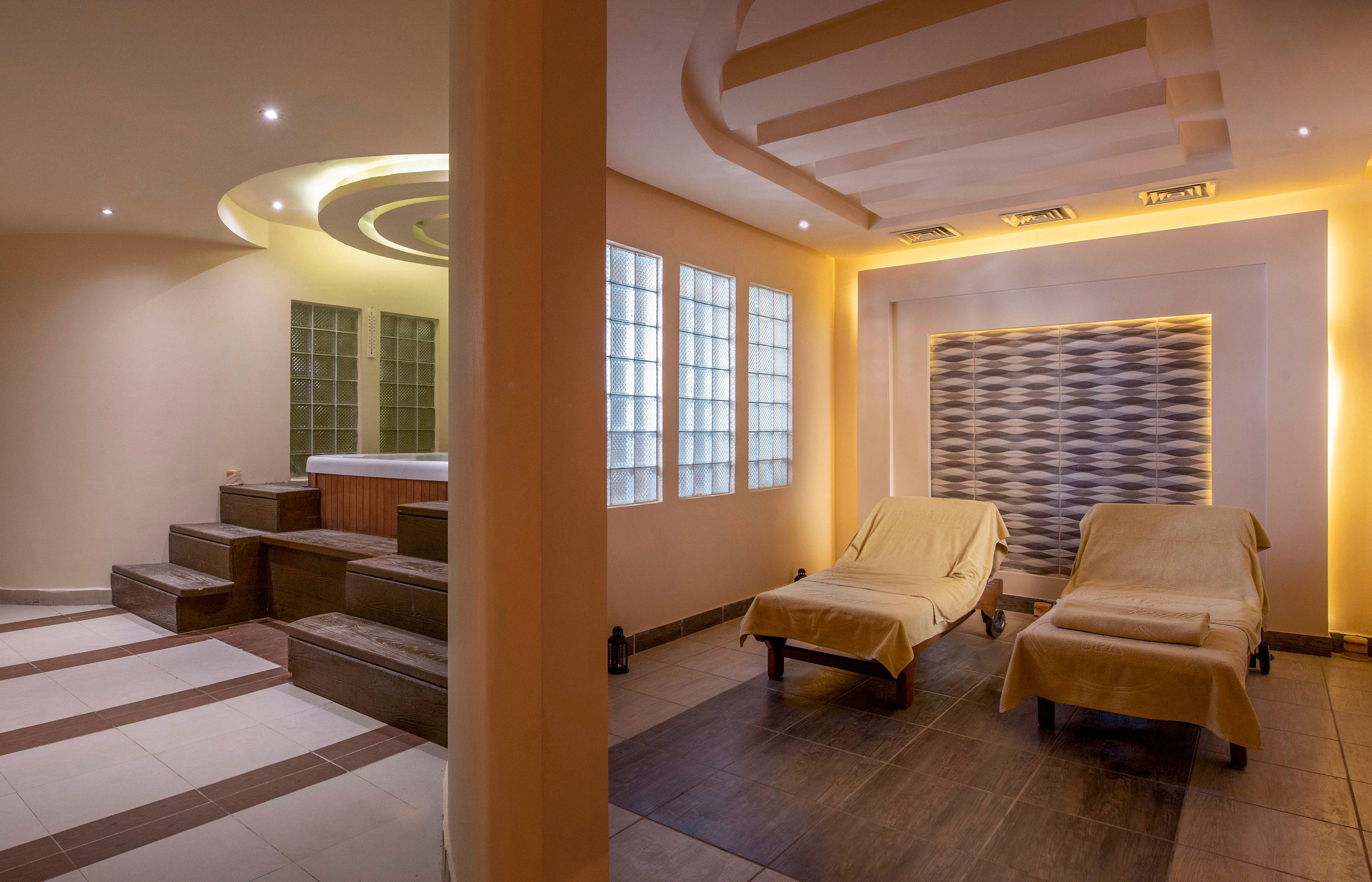 A serene spa lounge featuring a woman seated on a massage bed, dressed in a white robe. The room includes warm lighting, modern decor, and a jacuzzi area with steps. The ambiance is calm and inviting, ideal for relaxation and rejuvenation.