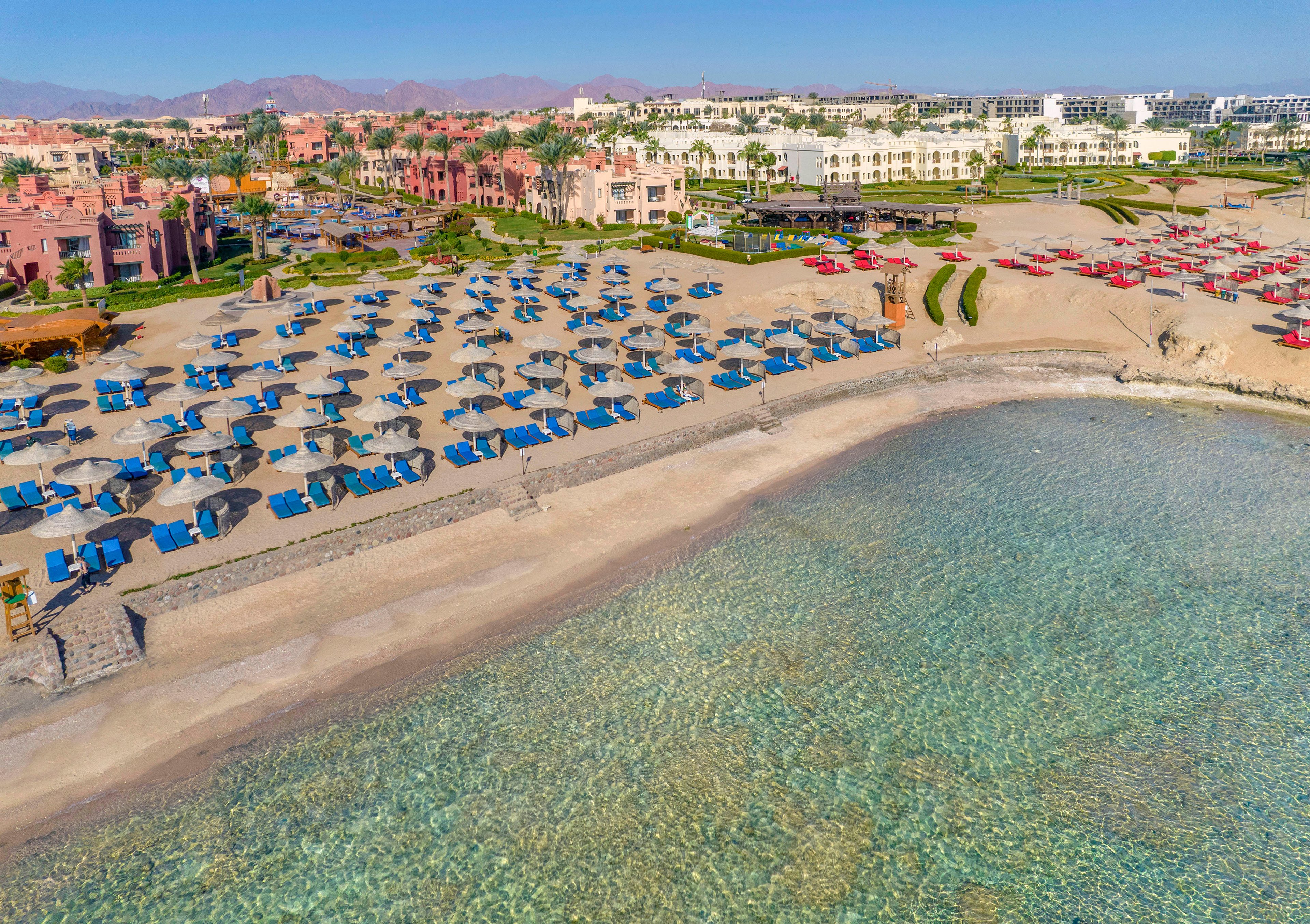 A scenic beachfront resort featuring rows of blue umbrellas and lounge chairs arranged neatly on the sandy shore. The crystal-clear water of the sea is visible in the foreground, with resort buildings and landscaped gardens in the background. The setting is tranquil and inviting, ideal for relaxation and vacation activities.