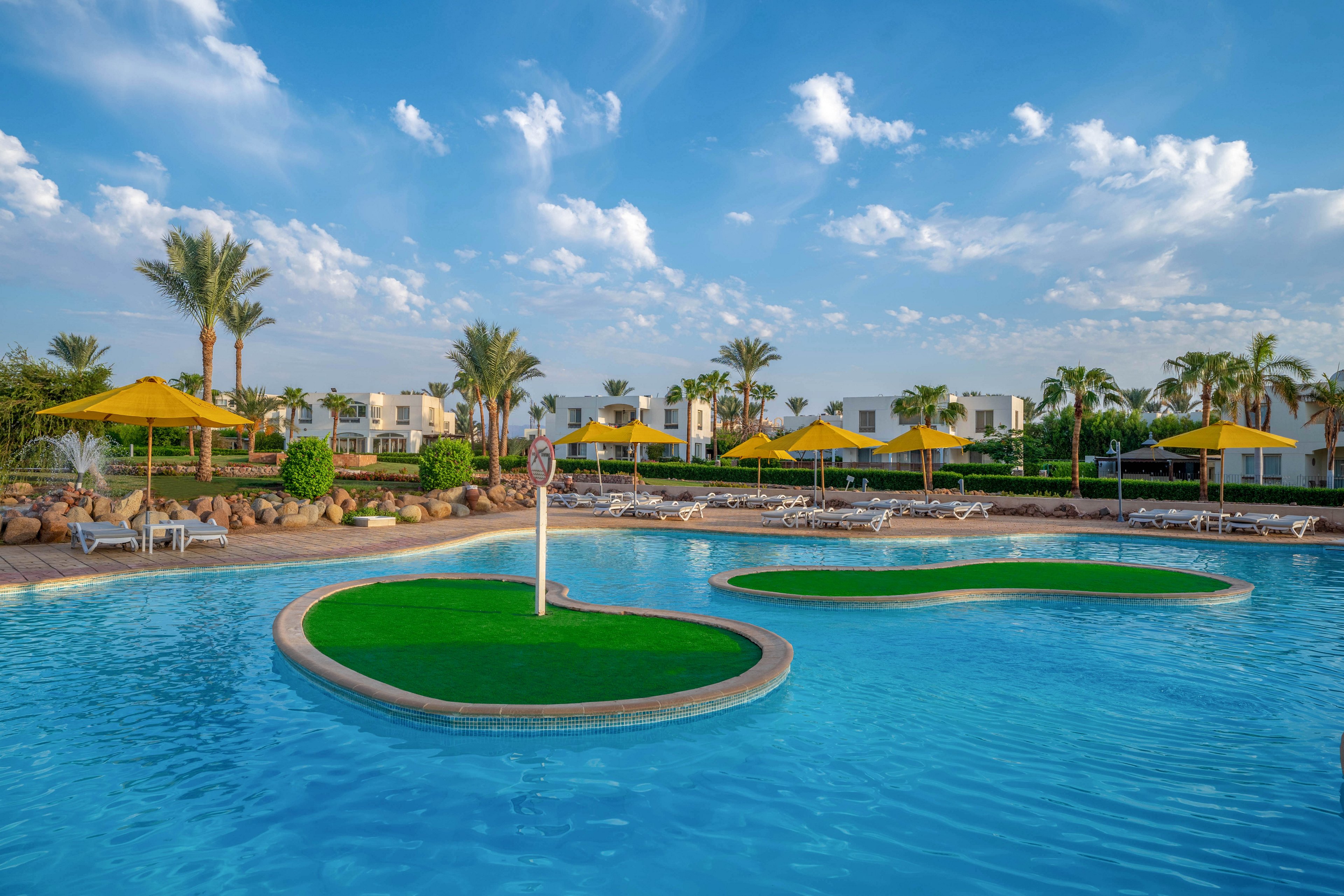 A large outdoor swimming pool is featured in the foreground, with two green artificial islands and a signpost. The setting includes palm trees, yellow umbrellas, and white lounge chairs, creating a relaxing resort atmosphere. Modern white buildings are visible in the background under a blue sky with scattered clouds. The scene is bright and tranquil, evoking a vacation mood.