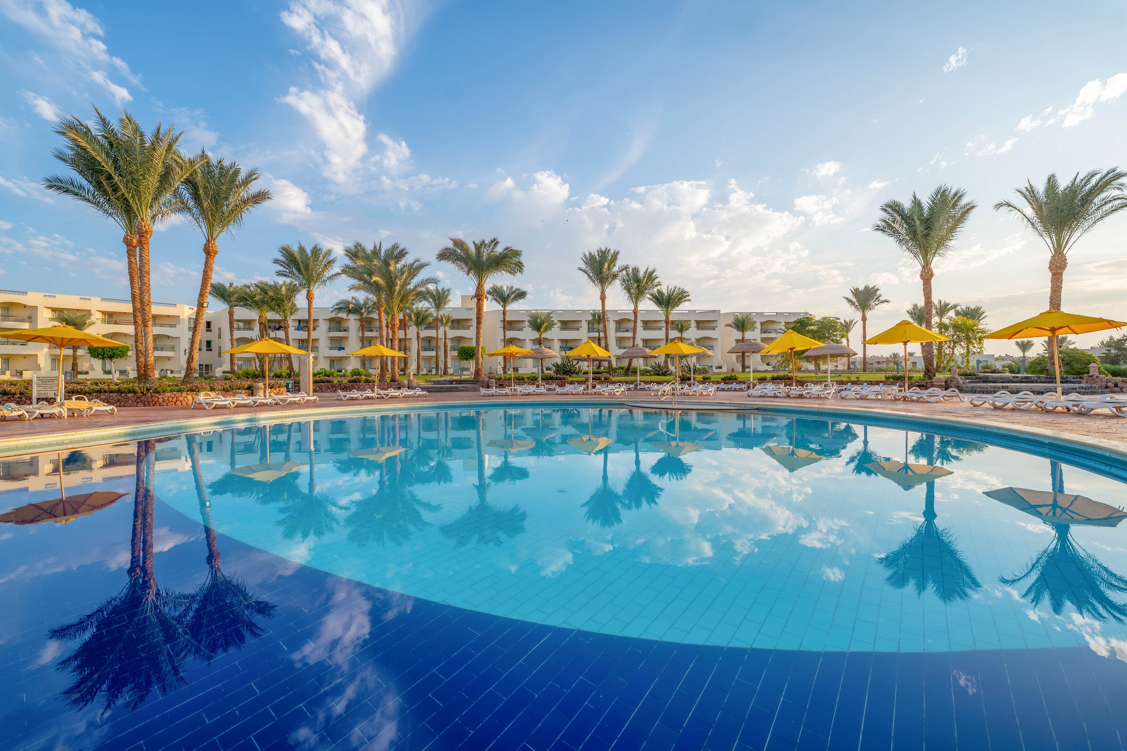 A large, circular swimming pool is the main focus, surrounded by tall palm trees and yellow umbrellas. The setting is a modern resort with white buildings in the background, under a bright blue sky with scattered clouds. The mood is tranquil and inviting, with reflections of the palm trees and sky visible in the water.
