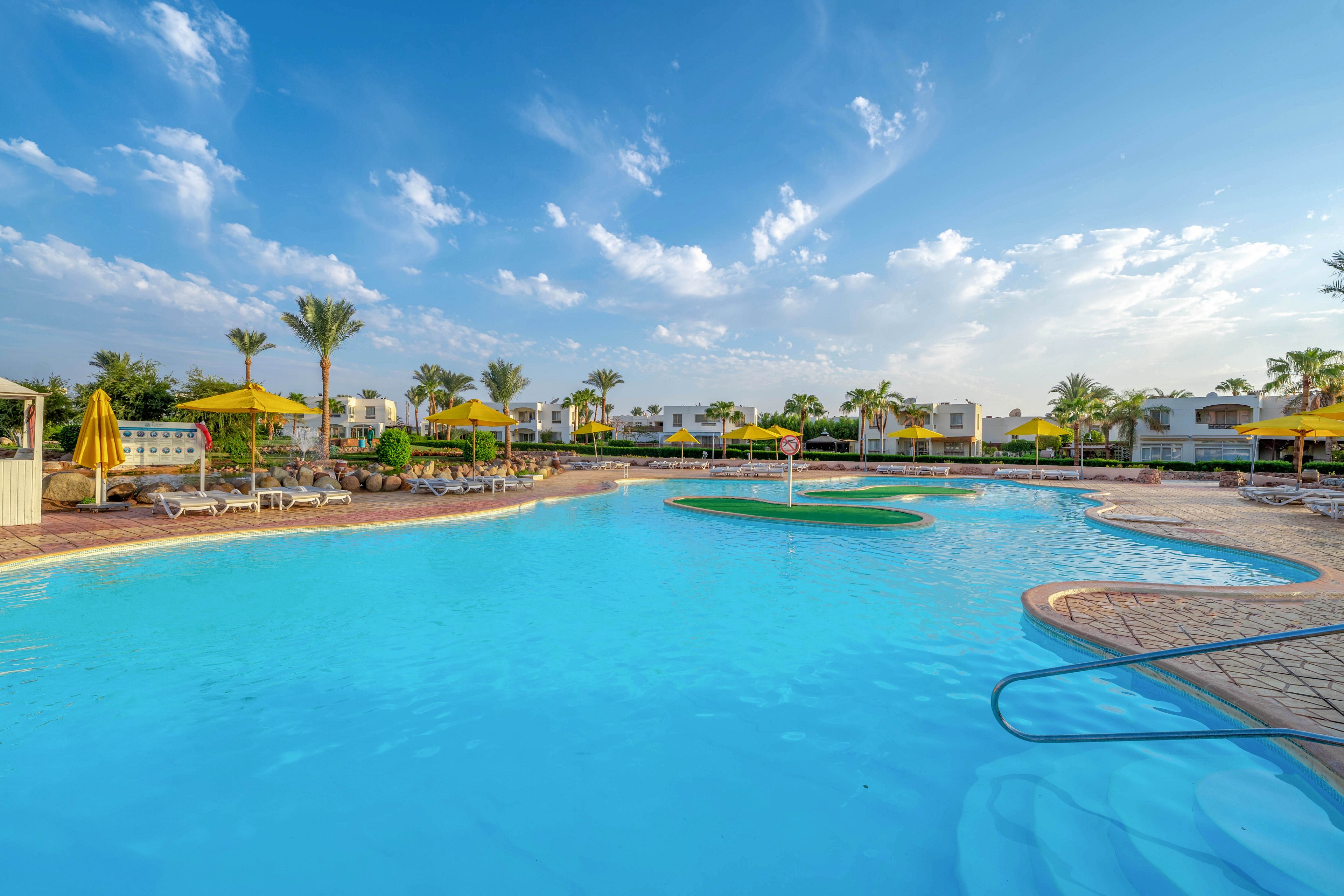 A large outdoor swimming pool is the main focus, surrounded by palm trees and yellow umbrellas. Lounge chairs line the poolside, and modern resort buildings are visible in the background. The scene is bright and tranquil, with clear blue skies and scattered clouds, evoking a relaxing vacation mood.