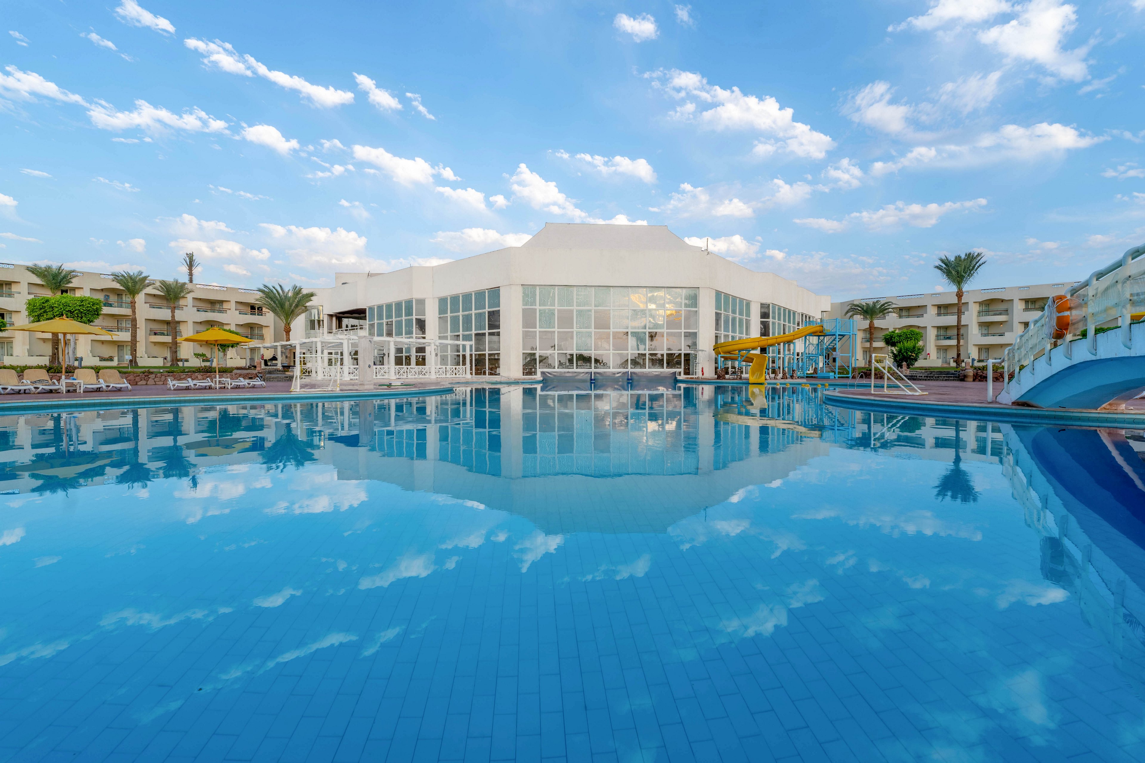 A large, contemporary hotel building is reflected in a spacious outdoor swimming pool. The setting features palm trees, lounge chairs, and water slides, suggesting a leisure and vacation atmosphere. The sky is bright with scattered clouds, enhancing the tranquil mood. The pool area is clean and inviting, with blue tones dominating the palette.