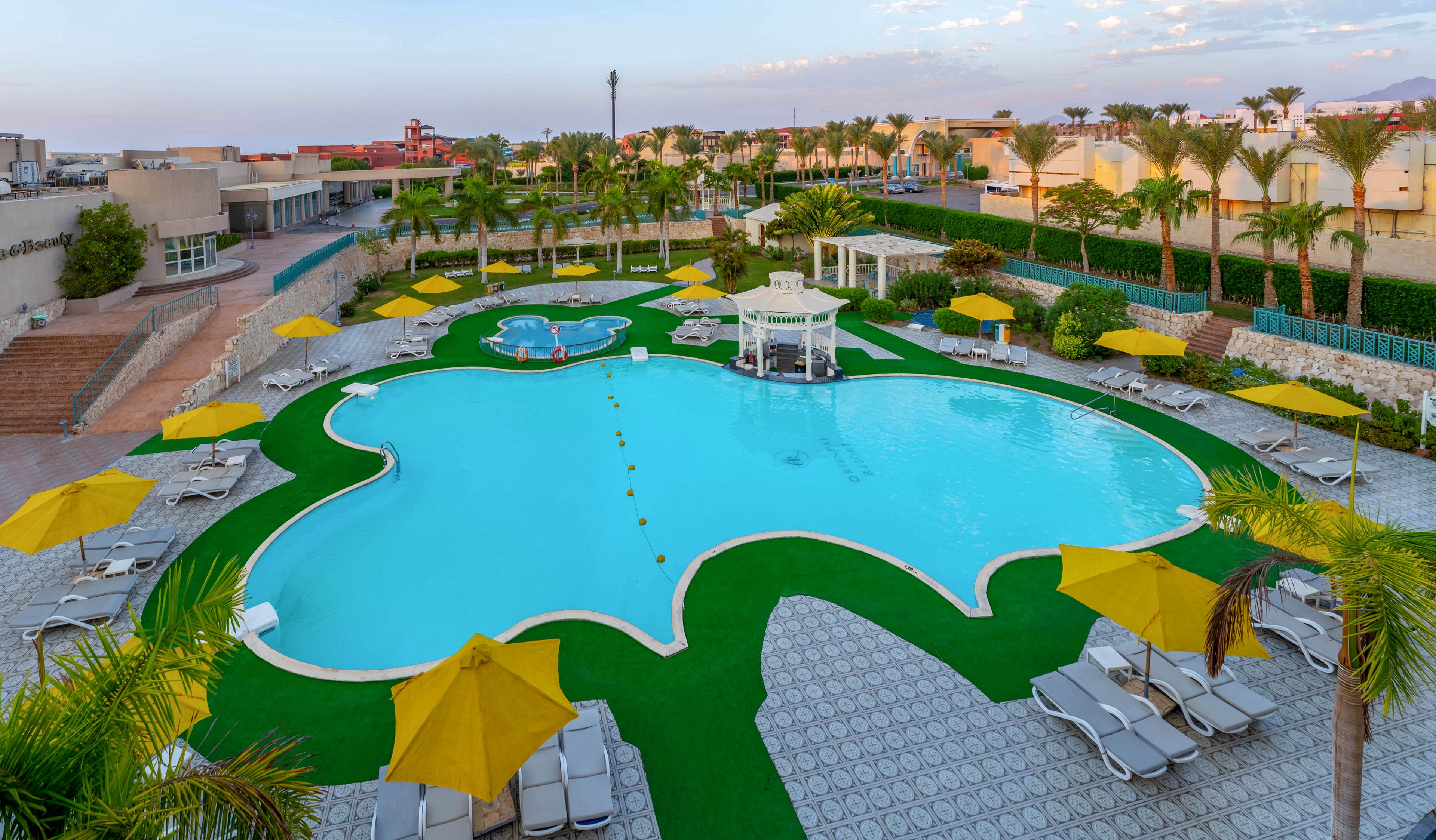A large, uniquely shaped swimming pool is the central feature of a resort outdoor area. Surrounding the pool are numerous lounge chairs and bright yellow umbrellas, set on patterned tiles and artificial grass. Palm trees and landscaped gardens frame the scene, with resort buildings visible in the background. The setting is calm and inviting, ideal for relaxation and leisure.