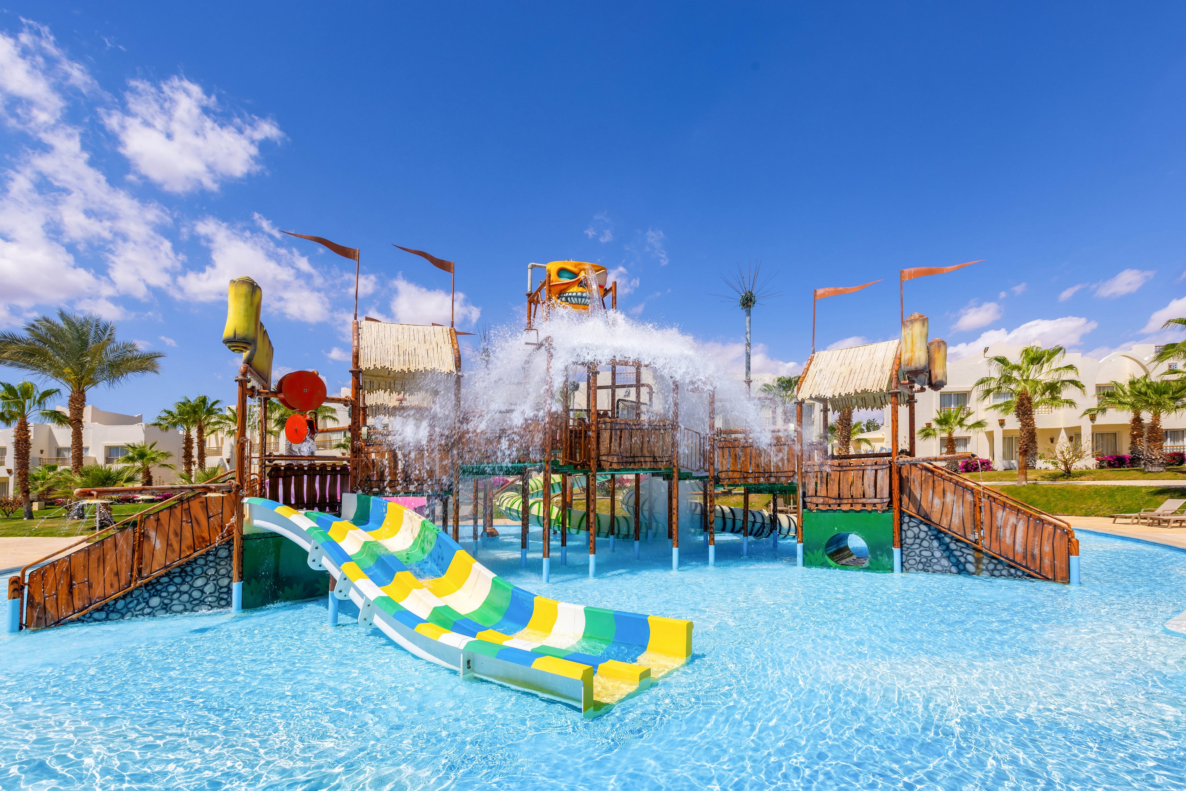 A vibrant water playground features multiple slides and interactive splash elements in a sunny outdoor setting. The structure includes green, yellow, and white striped slides leading into a shallow pool, with water jets and buckets creating playful splashes. Palm trees and resort buildings are visible in the background, enhancing the tropical atmosphere. The sky is bright blue with scattered clouds, contributing to a cheerful and inviting mood.