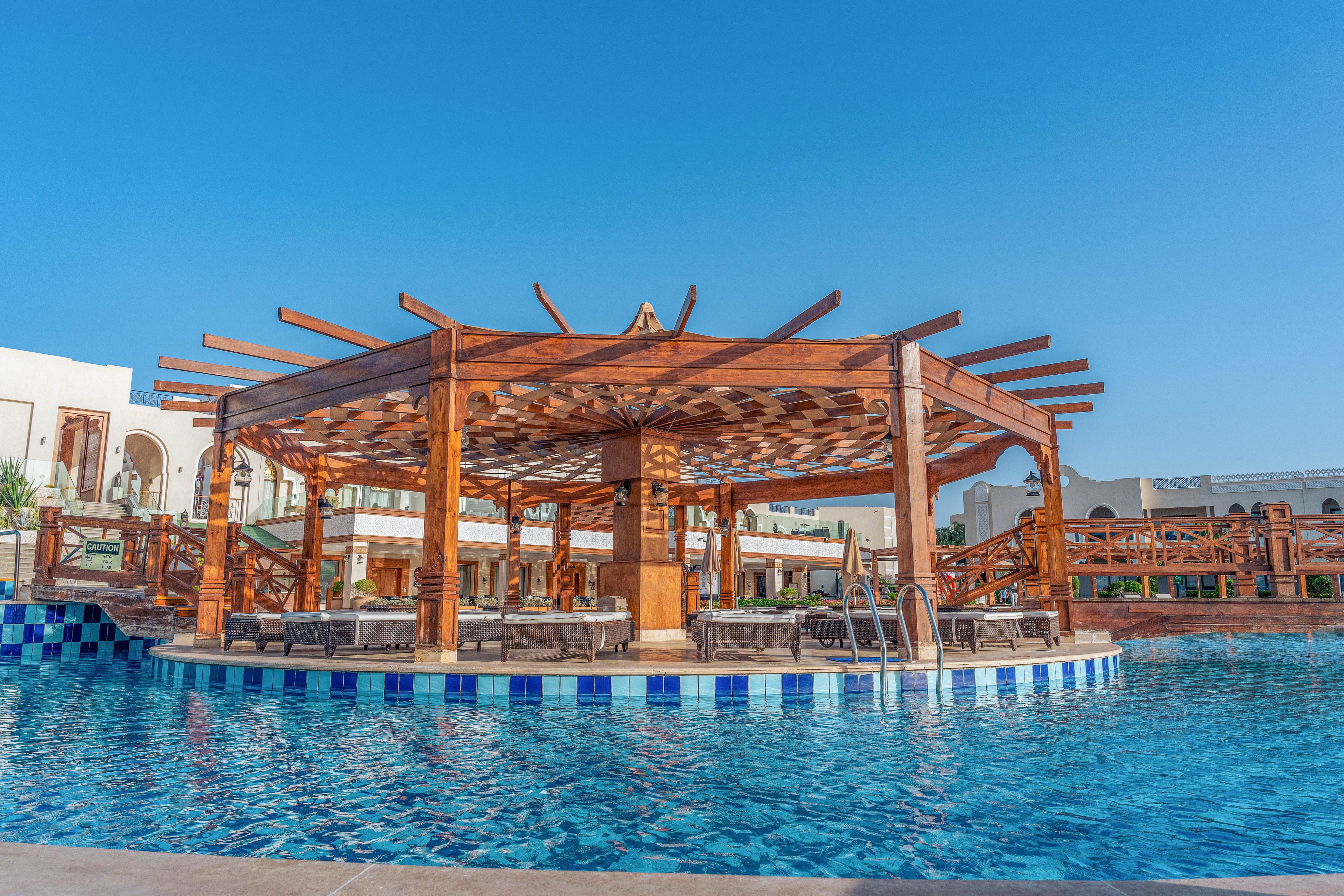 A large resort swimming pool features a circular wooden pavilion with shaded lounge chairs arranged around it. The structure sits in the middle of the blue-tiled pool, accessible by metal ladders. Surrounding buildings with balconies and warm-toned facades frame the scene under a clear blue sky. The overall atmosphere suggests a relaxing vacation destination with inviting water and comfortable seating.