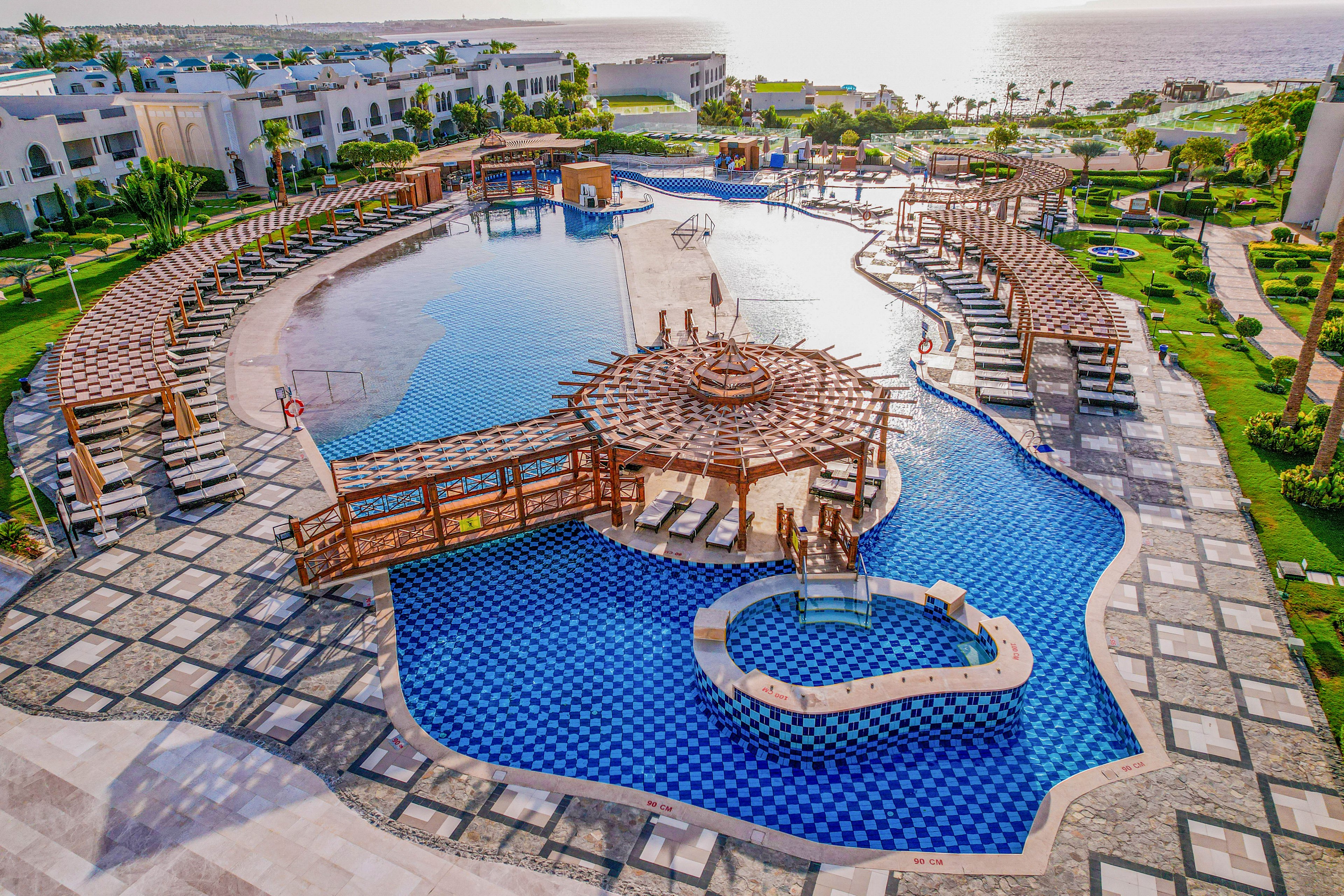 A large resort swimming pool with blue mosaic tiles is surrounded by sun loungers and wooden pergolas. Curved bridges and a central circular deck create an intricate pool layout. White resort buildings and manicured gardens frame the scene. In the distance, the ocean glows under the bright sunlight, enhancing the tropical vacation atmosphere.