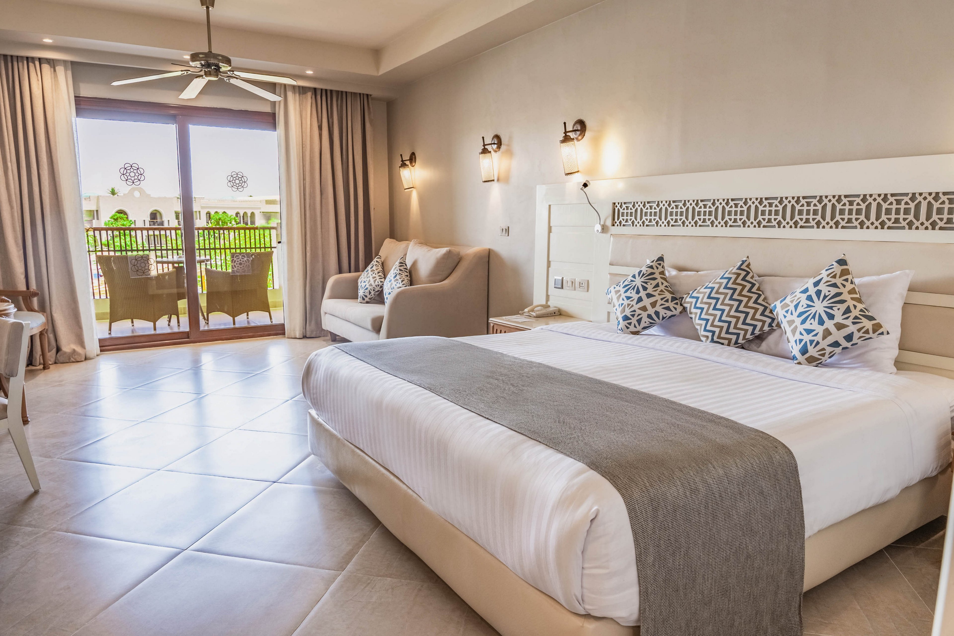 A spacious, contemporary hotel bedroom features a large neatly made bed with a gray throw and patterned pillows. The room includes a comfortable sofa, ceiling fan, and light tiled flooring that enhances the airy feel. Floor-to-ceiling curtains frame glass doors leading to a balcony with outdoor seating and greenery visible outside. Neutral tones and soft lighting create a calm, inviting atmosphere ideal for relaxation.