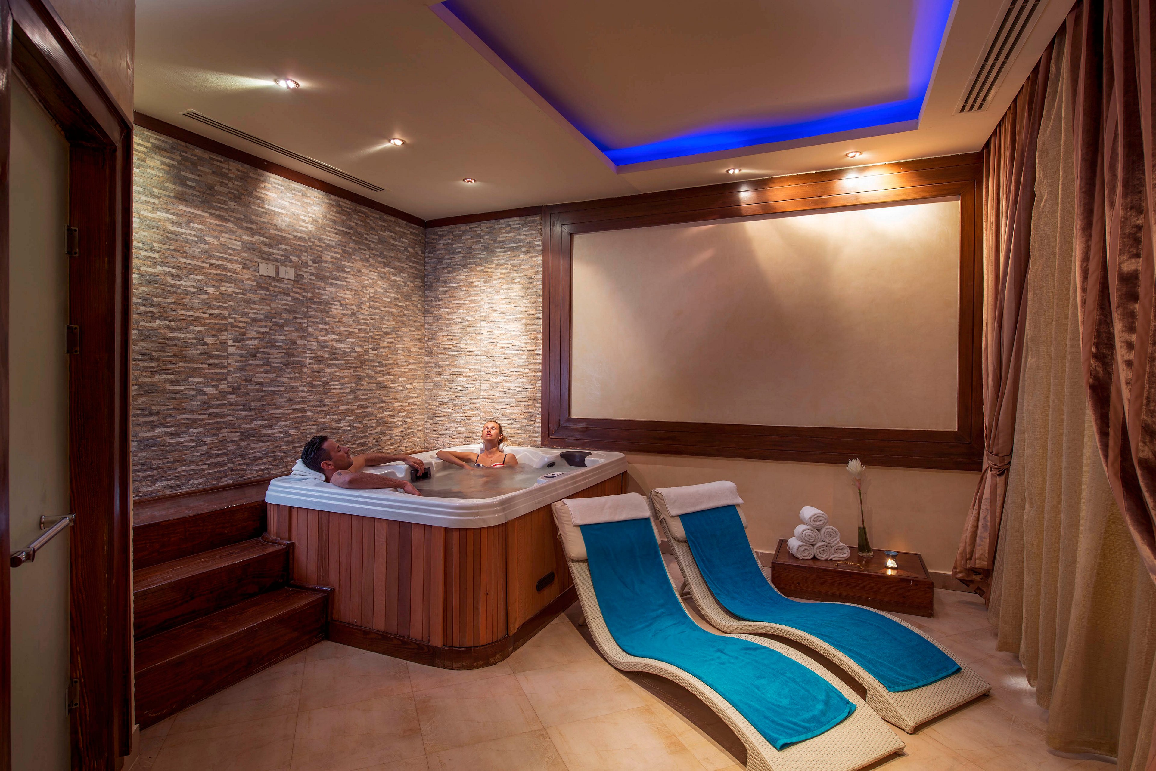 A couple relaxes together in a built-in jacuzzi tub inside a luxurious spa room. The hot tub is set into a wooden platform with stone-textured walls and soft, warm lighting. Two modern lounge chairs with bright blue cushions sit in the foreground on a tiled floor. The ceiling features recessed lighting with a blue accent glow, and curtains and folded towels add to the tranquil atmosphere.