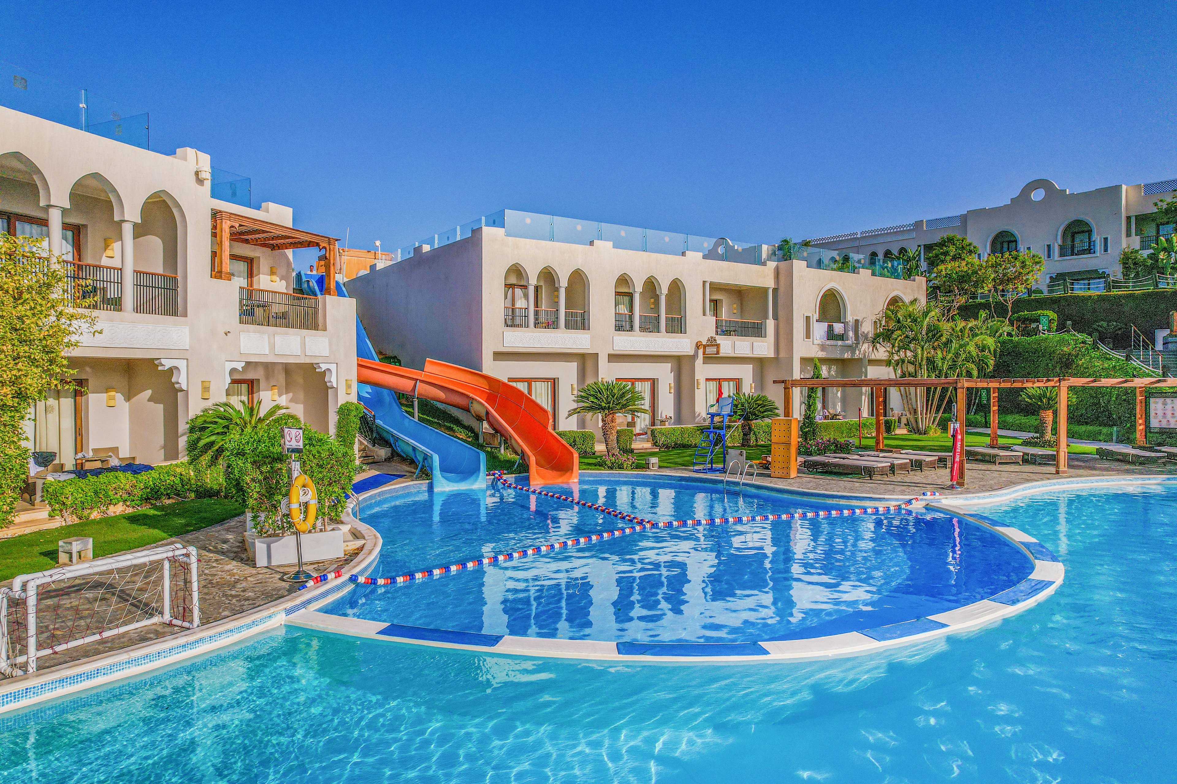 A bright resort pool area features curved blue water and multiple colorful waterslides leading into it. The surrounding buildings have white facades with arches and balconies overlooking the pool. Lush green plants and palm trees add a tropical feel to the scene. Clear blue sky and vivid colors create a cheerful, family-friendly vacation atmosphere.