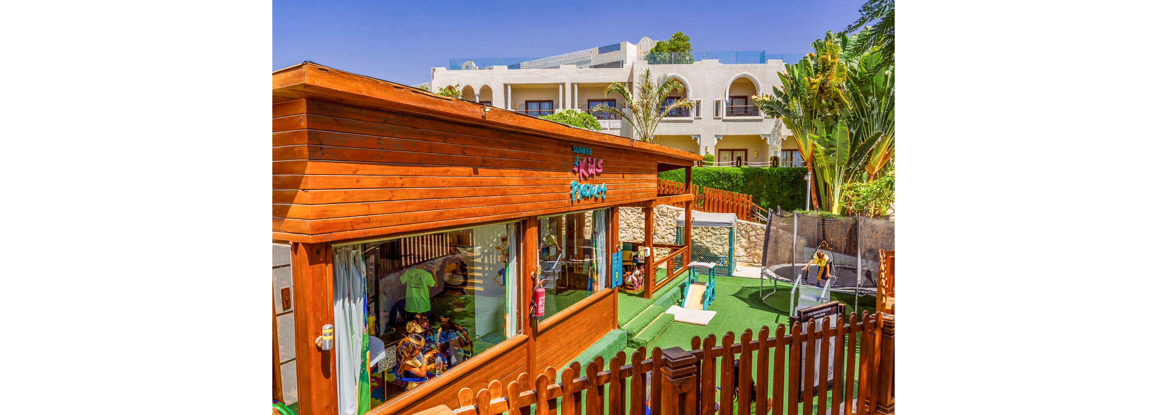 A vibrant wooden kids club building stands in an outdoor play area at a sunny resort. Children and staff are visible inside and around the structure, engaging in activities. The playground features artificial grass, play equipment, and a fenced boundary for safety. In the background, white resort buildings and lush tropical plants create a bright holiday atmosphere.